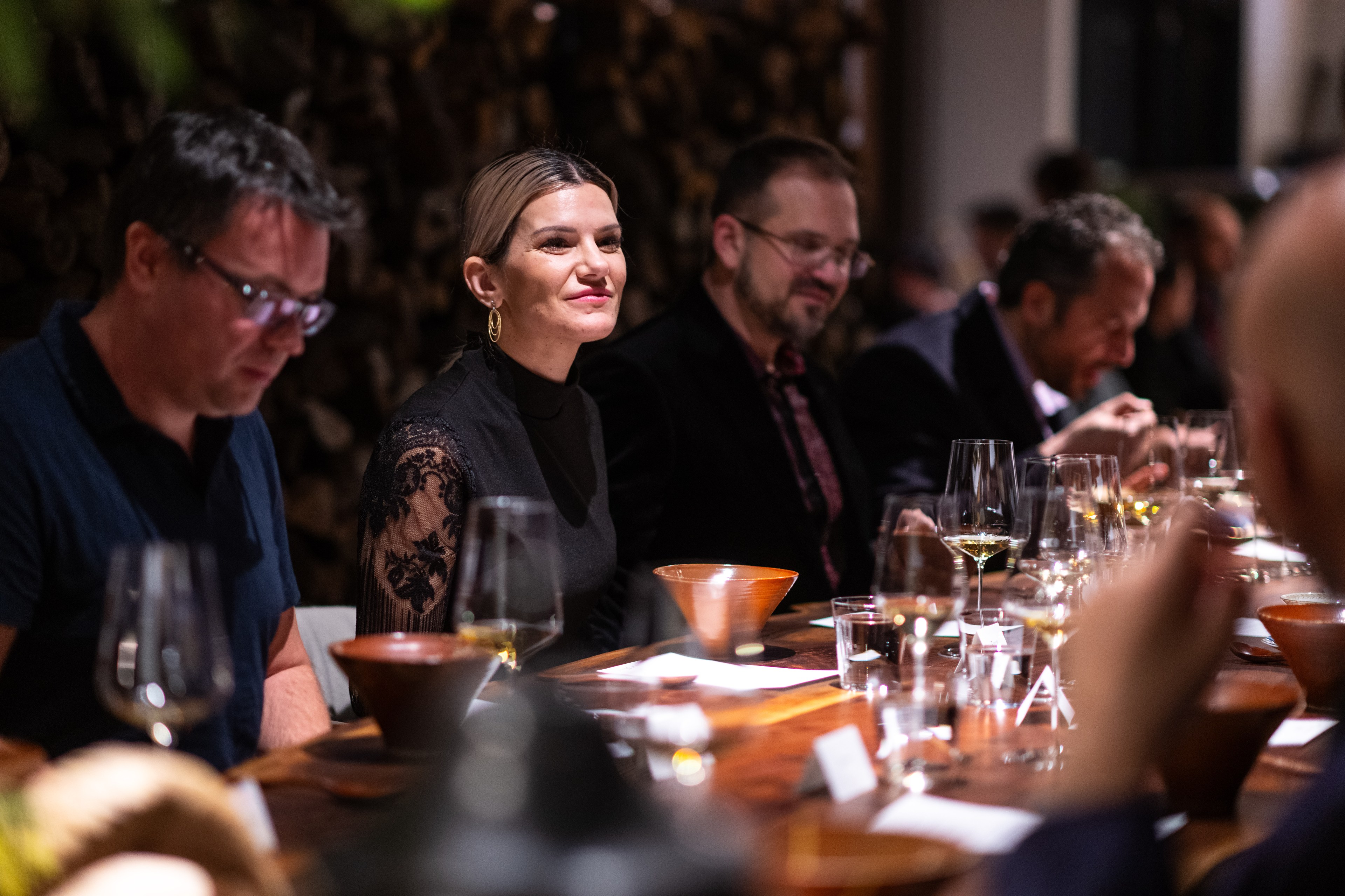 A group of people sit around a wooden table, engaged in conversation, with wine glasses and bowls in front of them. A woman in a black dress is smiling.