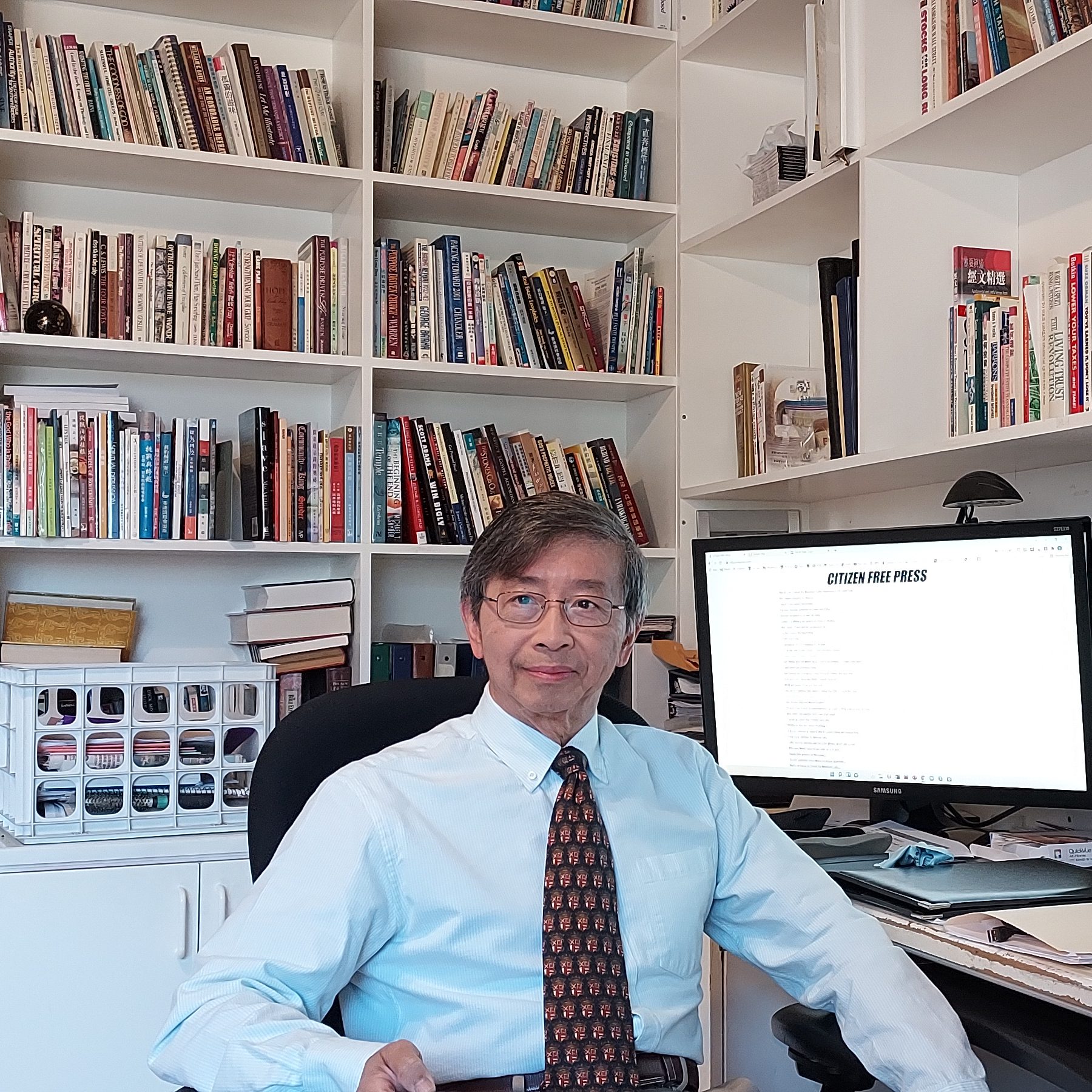 A person in a light blue shirt and patterned tie sits at a desk. Behind them are white bookshelves filled with various books. A computer monitor displays text.