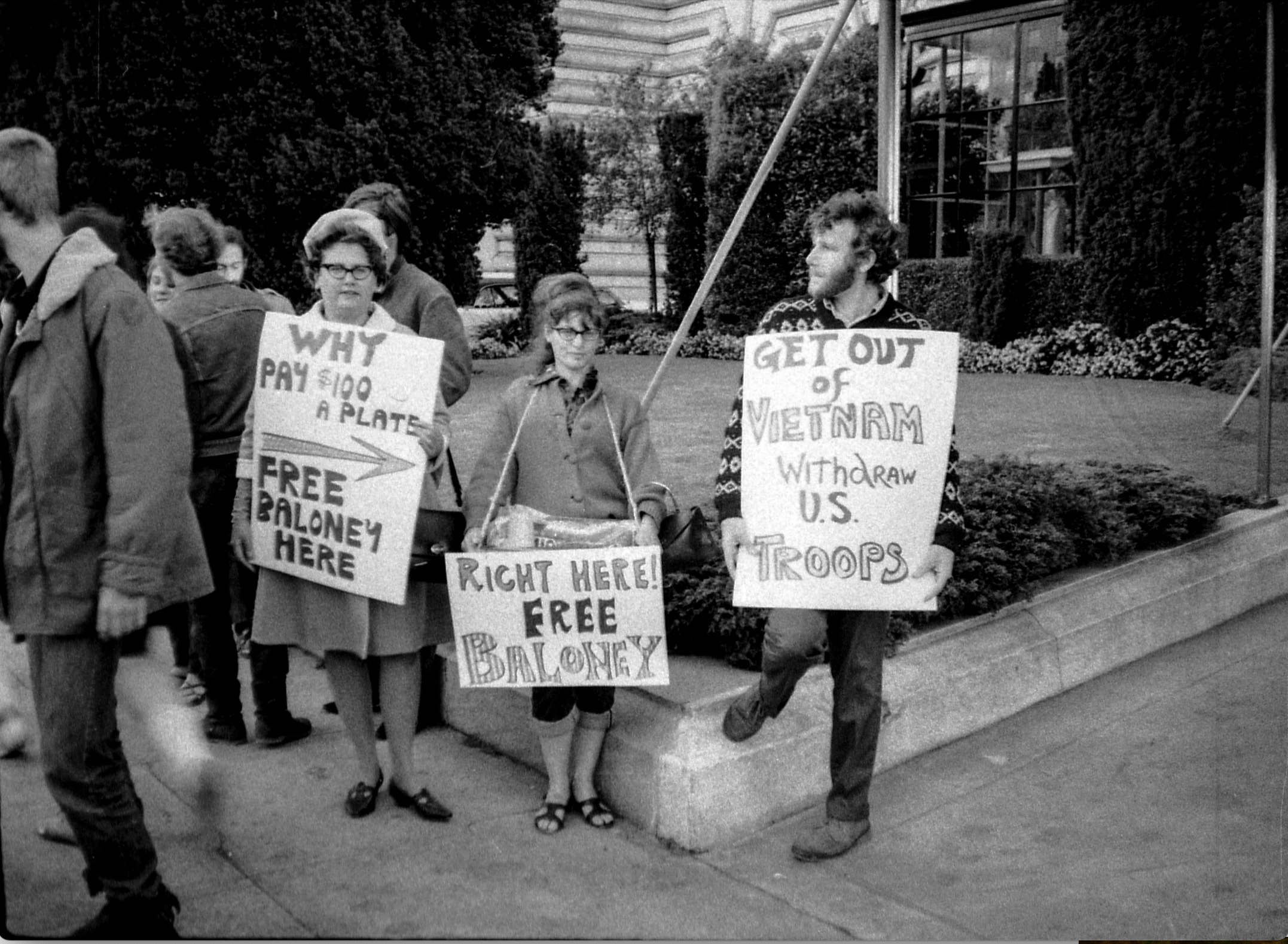 Three people hold protest signs, one reads &quot;Get Out of Vietnam,&quot; another offers &quot;Free Baloney,&quot; and others express dissent about expensive meals.