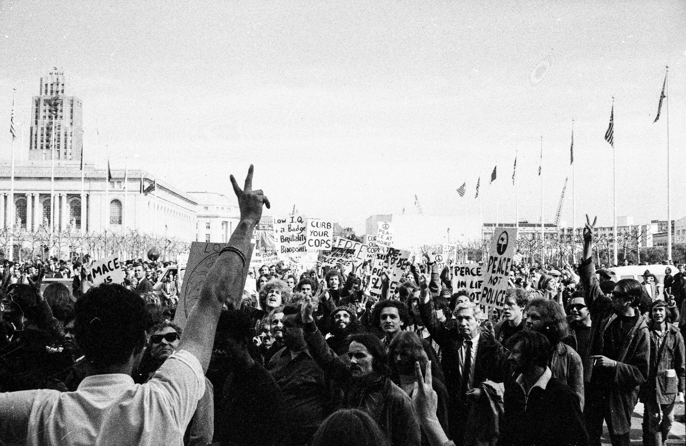 A large crowd gathers for a protest, holding signs with messages about peace and police reform. People raise their hands, and flags fly in the background.