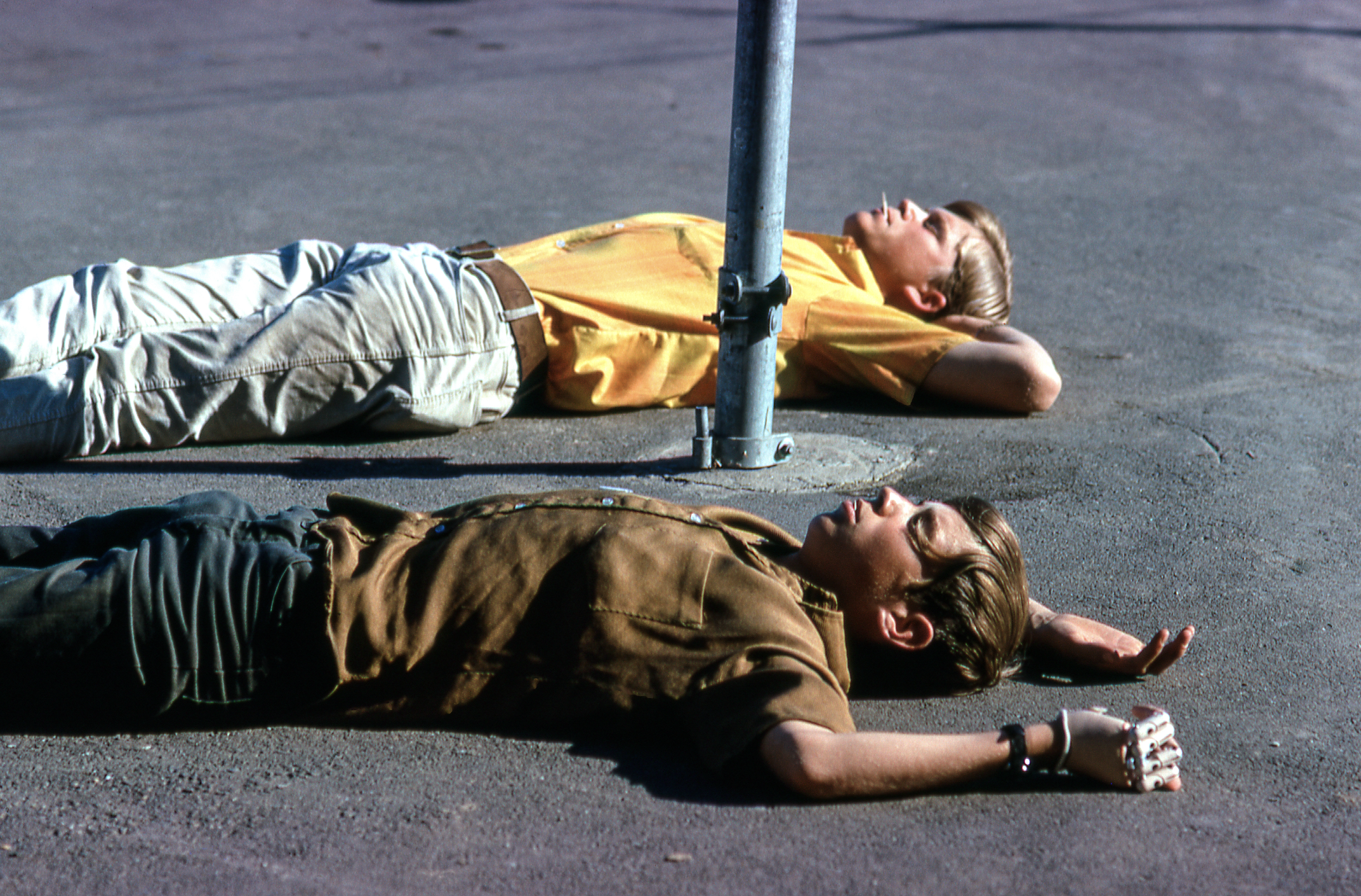 Two boys lie on the ground, their heads near a metal pole. One wears a yellow shirt, the other brown, both with arms behind their heads on a sunny day.