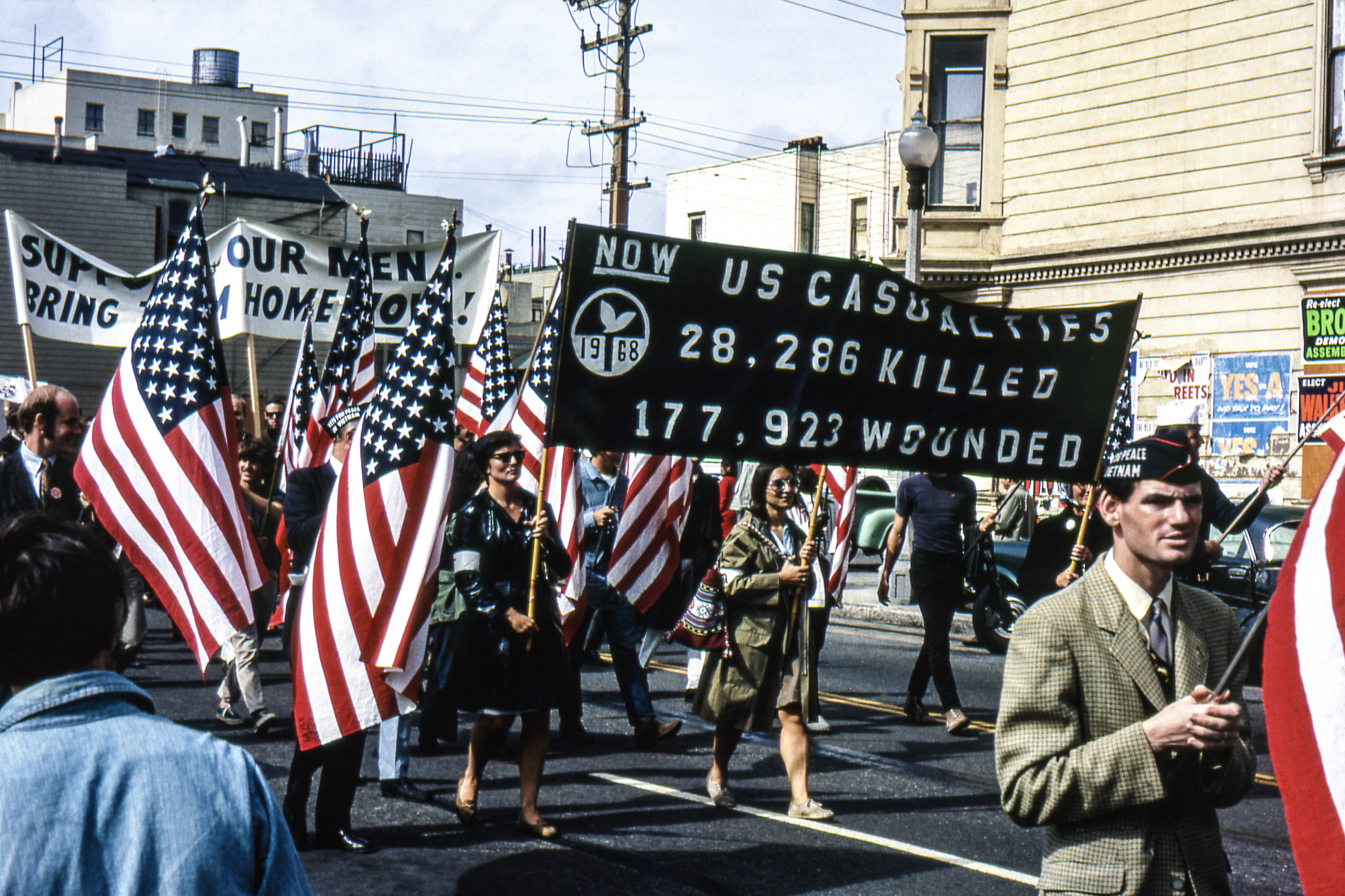 A protest march with people holding American flags and signs. One sign lists U.S. casualties: 28,286 killed and 177,923 wounded. Earth tones and mid-century urban backdrop.