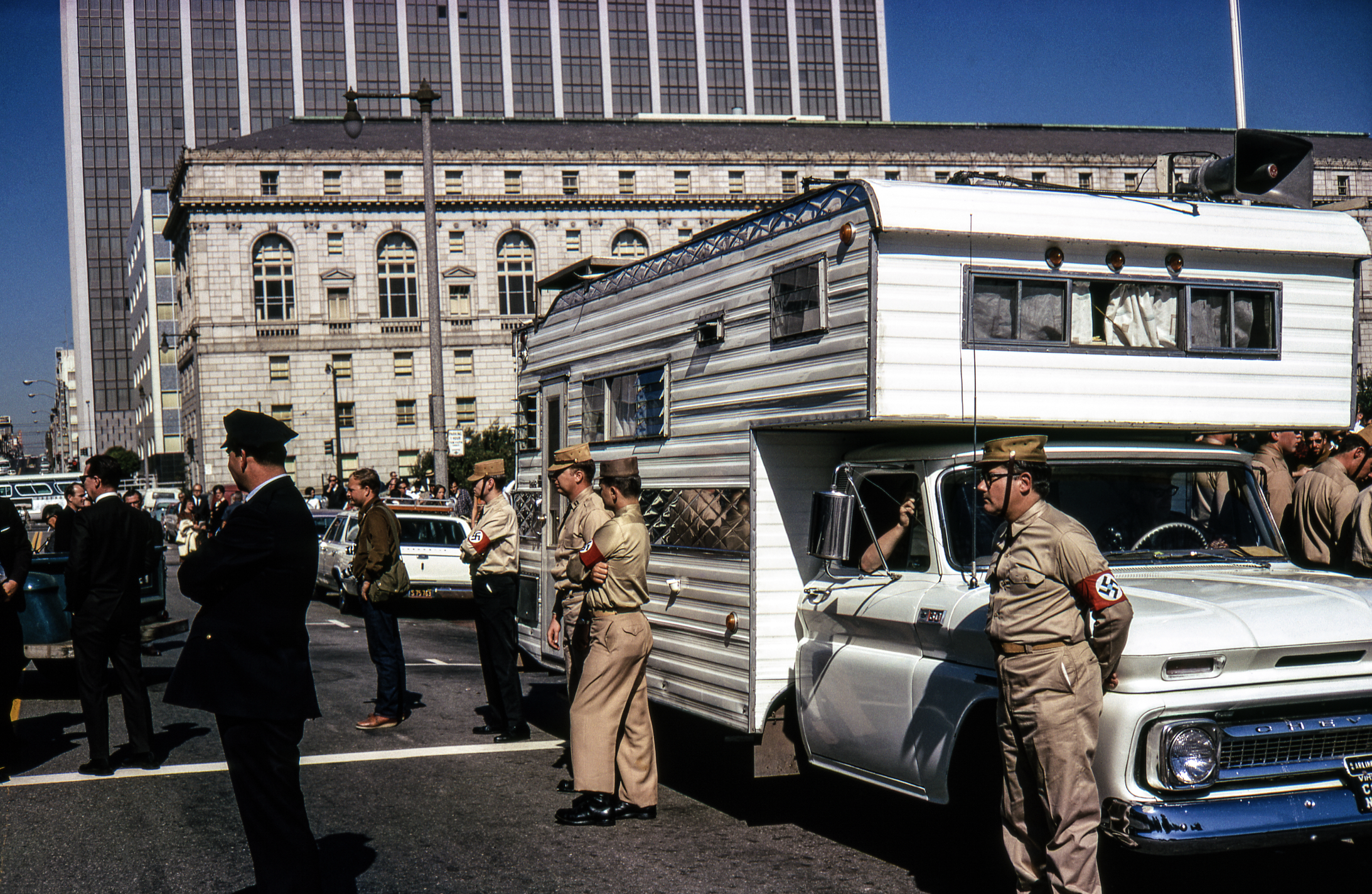 People in military uniforms, some with armbands, stand beside a white camper in an urban setting, surrounded by onlookers and tall buildings.