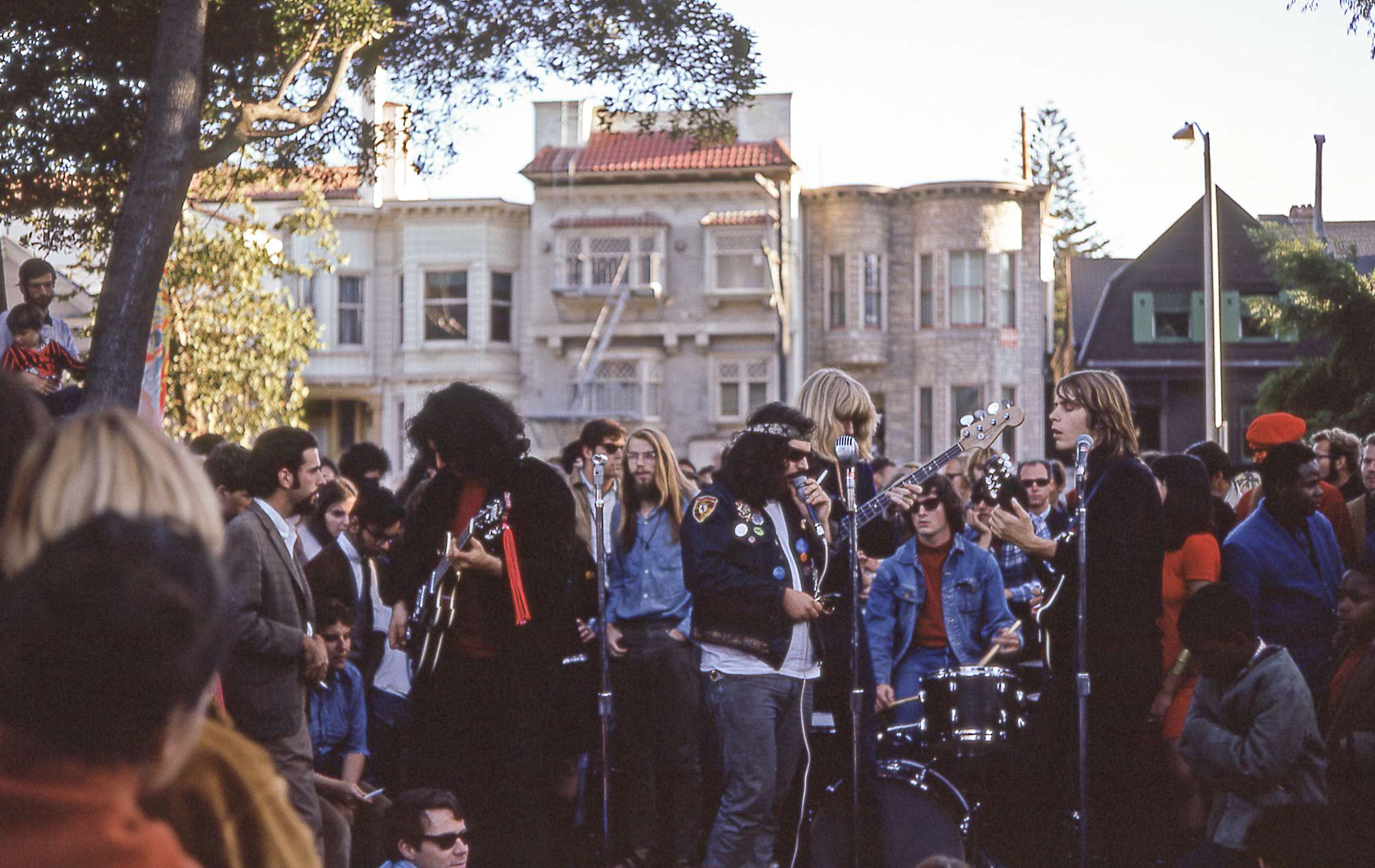 A band performs outdoors with guitars and drums, surrounded by a crowd. The background features trees and residential buildings under a clear sky.