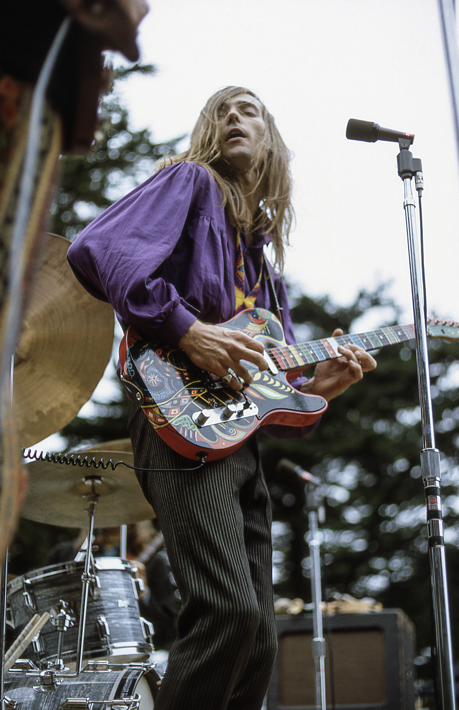 A musician with long hair in a vibrant purple shirt passionately plays an intricately painted guitar on stage. Drums and mics are visible in the background.