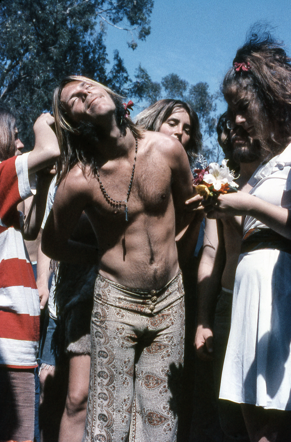 A shirtless man with long hair and a beaded necklace leans back among a group of people. He wears paisley pants, and the scene has a relaxed, bohemian vibe.