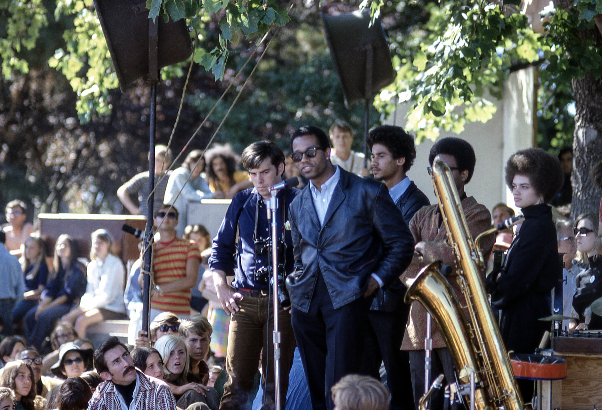 A man in a leather jacket speaks at a microphone outside, surrounded by a diverse group of people. A large crowd sits and stands nearby, listening intently.