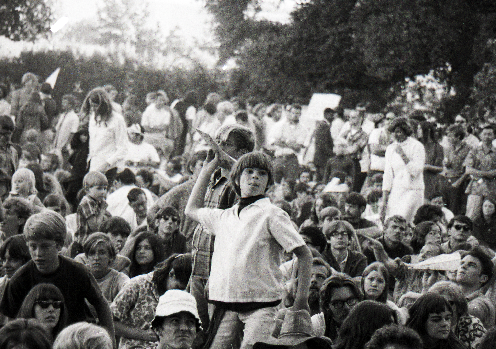 The image shows a large, diverse crowd outdoors, with people sitting and standing closely together. A child in the center looks engaged, holding a flag.