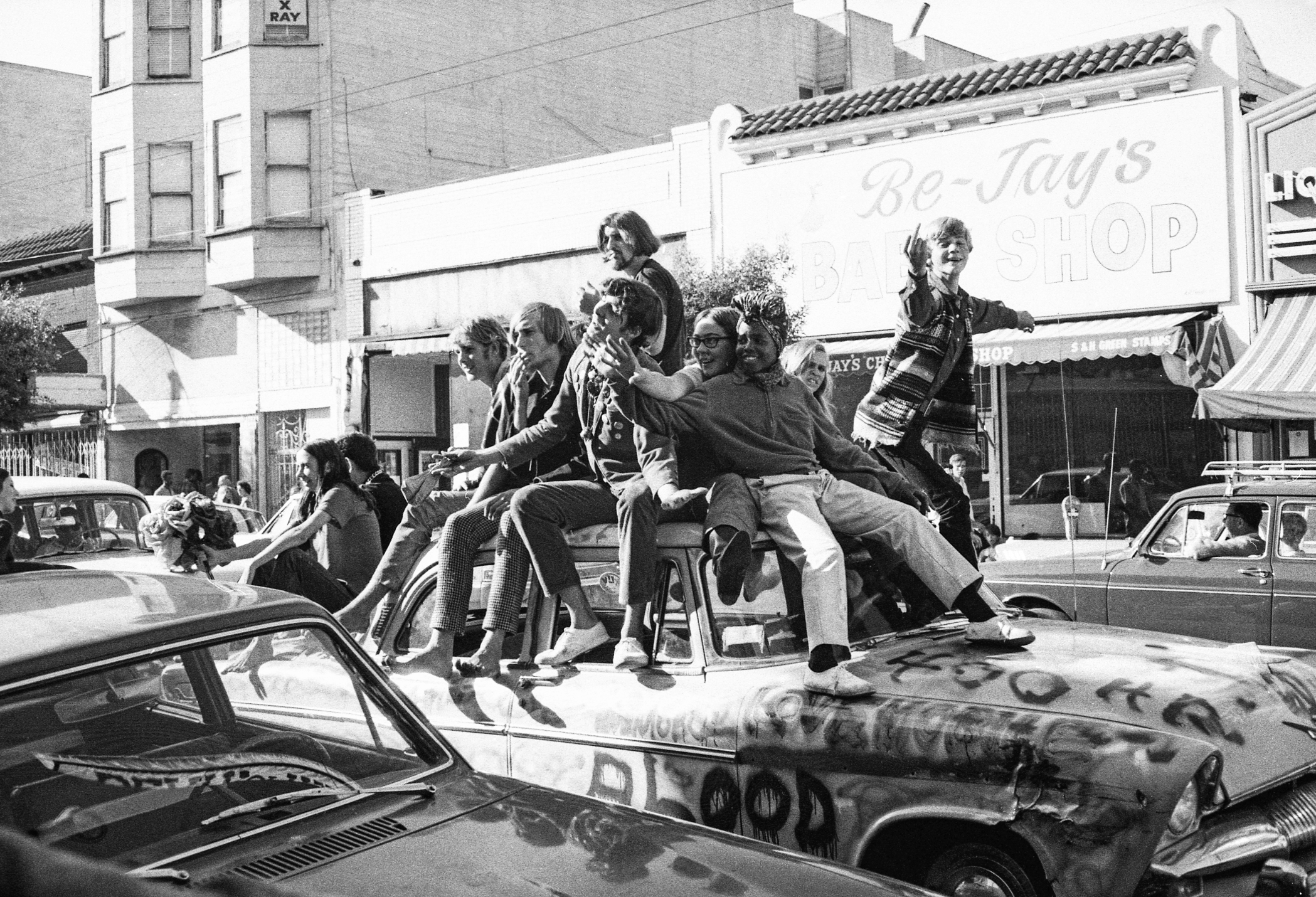 A group of people dressed in 1960s attire are sitting and standing on a graffiti-covered car, parked in front of a bakery, in a lively street scene.