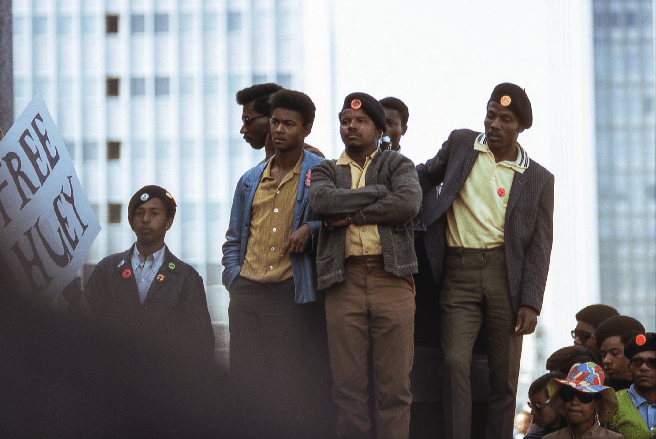 A group of men in yellow shirts and berets with badges stand in an urban setting. One holds a &quot;Free Huey&quot; sign. They appear serious and attentive.