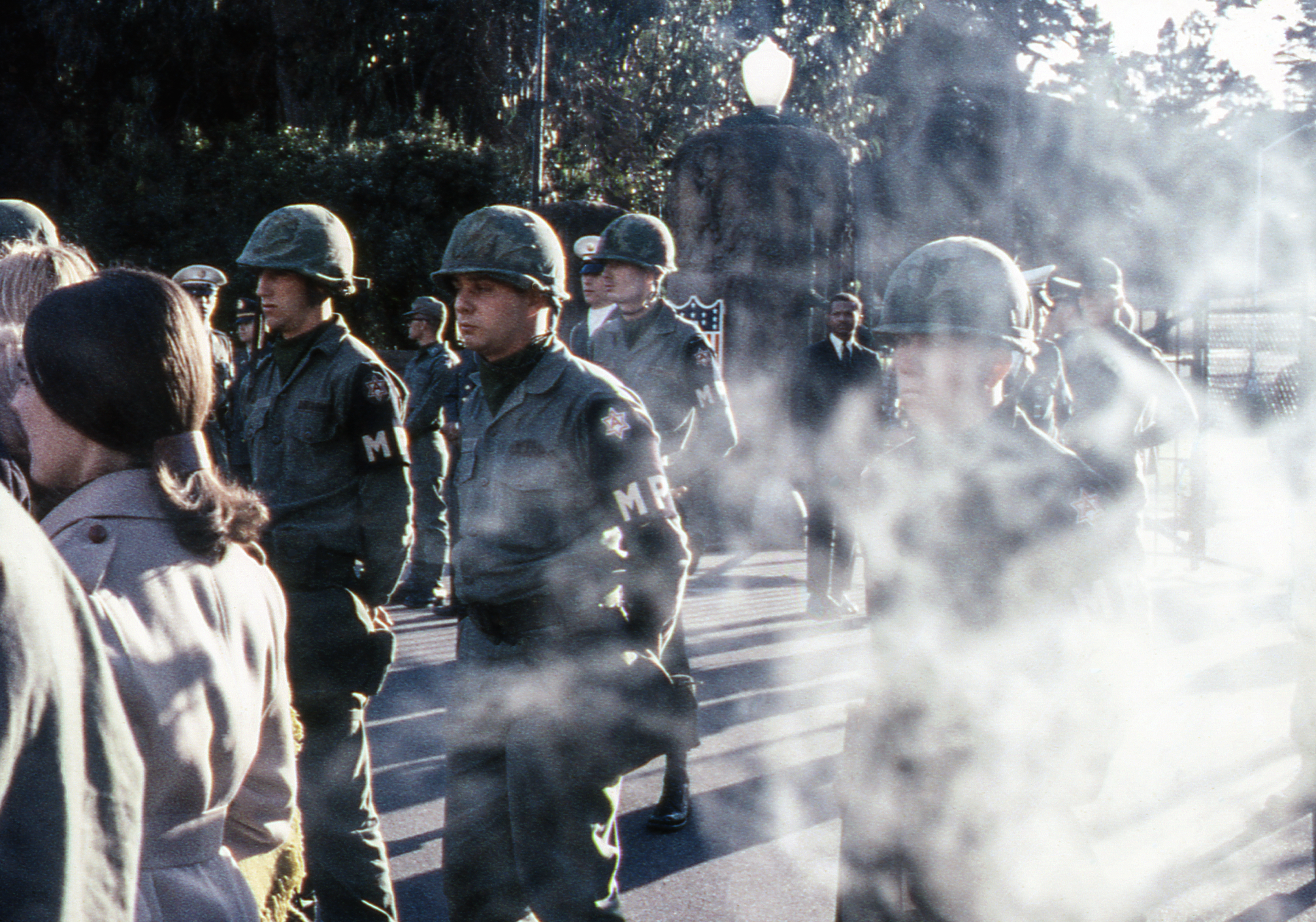 The image shows military police officers in uniform, wearing helmets, marching in formation. There is some smoke or fog obscuring part of the scene.