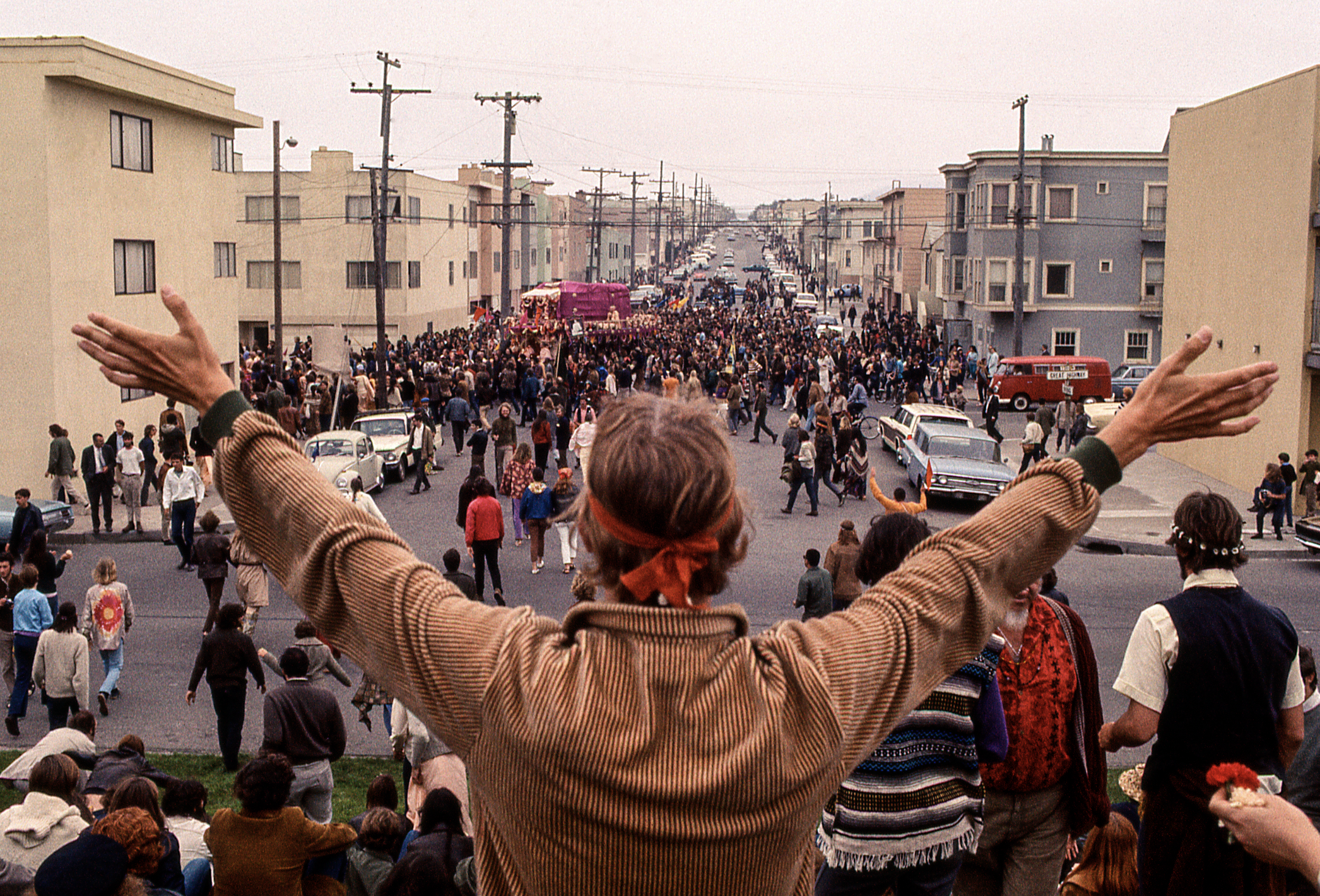 A large crowd fills a street with a person in the foreground raising their arms. People are walking, and some vehicles are parked along the sides.