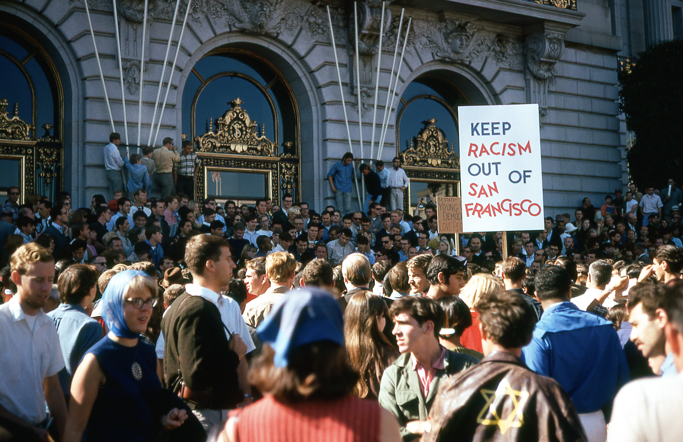 A large crowd gathers in front of an ornate building. A prominent sign reads &quot;Keep Racism Out of San Francisco,&quot; indicating a protest or demonstration.