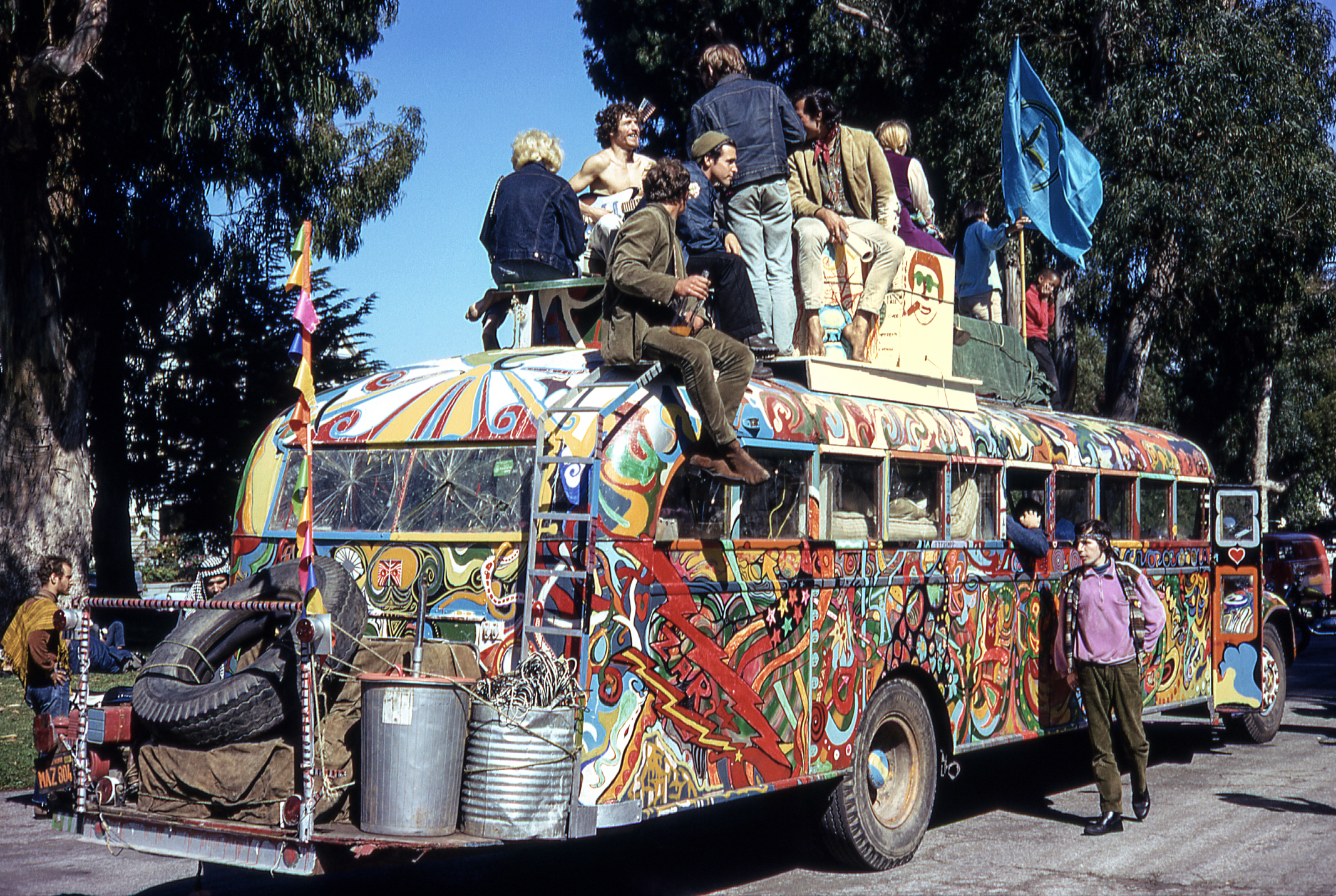 A vibrantly painted bus with psychedelic patterns has several people sitting on the roof. They are surrounded by trees and there's a blue flag waving.