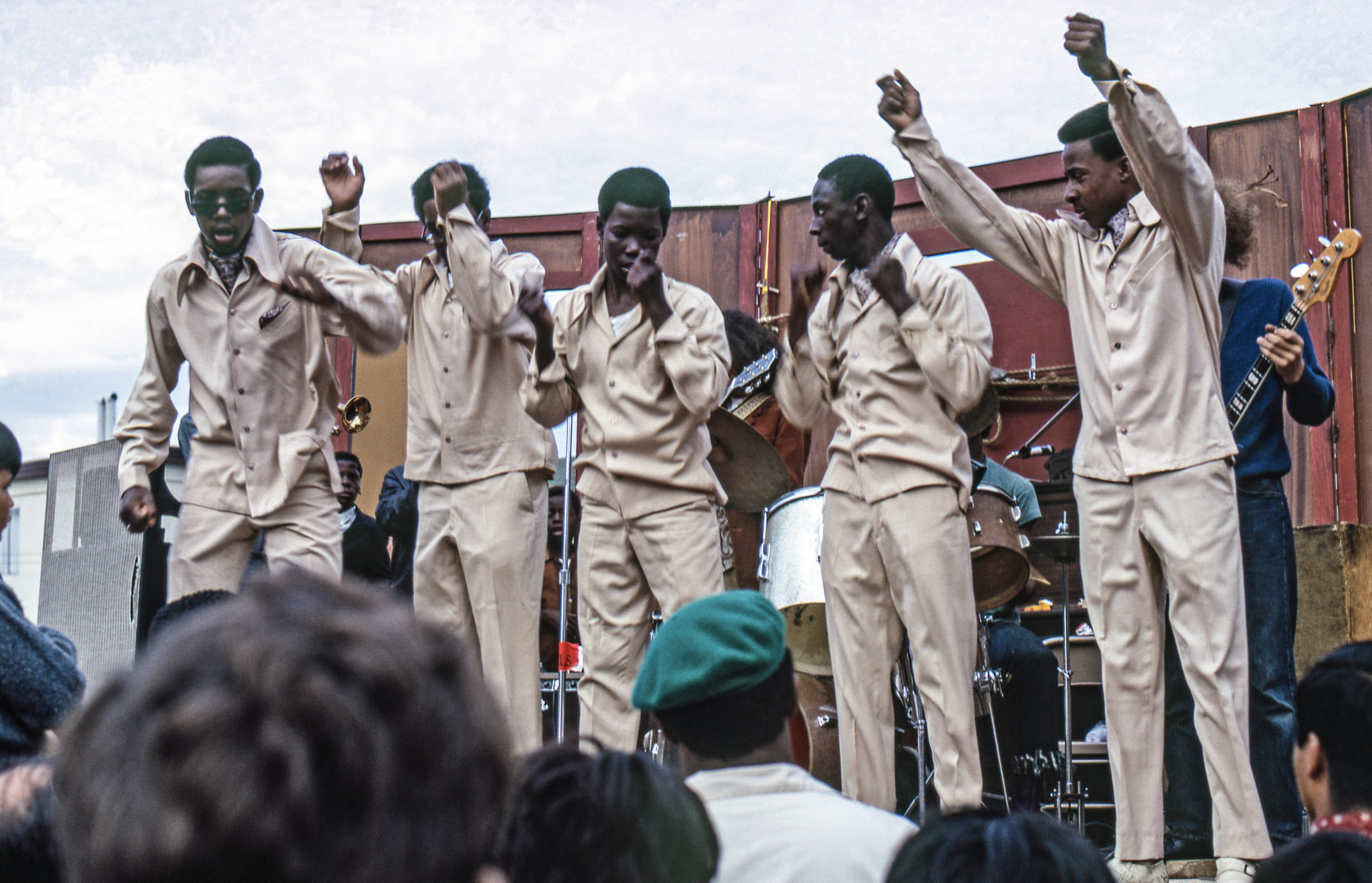 Five men in beige outfits perform energetically on an outdoor stage with musical instruments, surrounded by an audience. They look lively and animated.