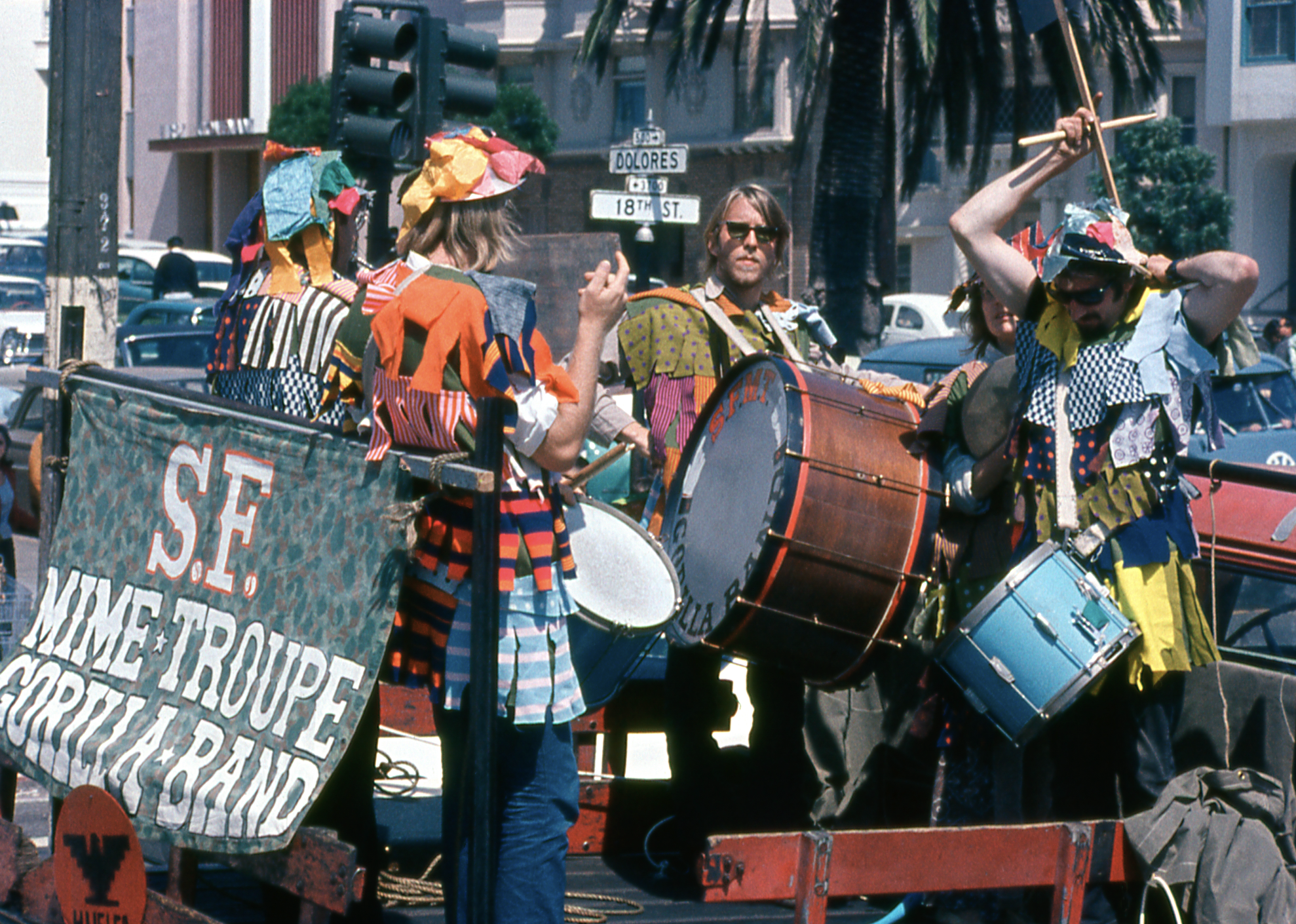 A colorful band with patchwork costumes plays drums on a street corner. A banner reads &quot;S.F. Mime Troupe Gorilla Band,&quot; with vehicles and buildings in the background.