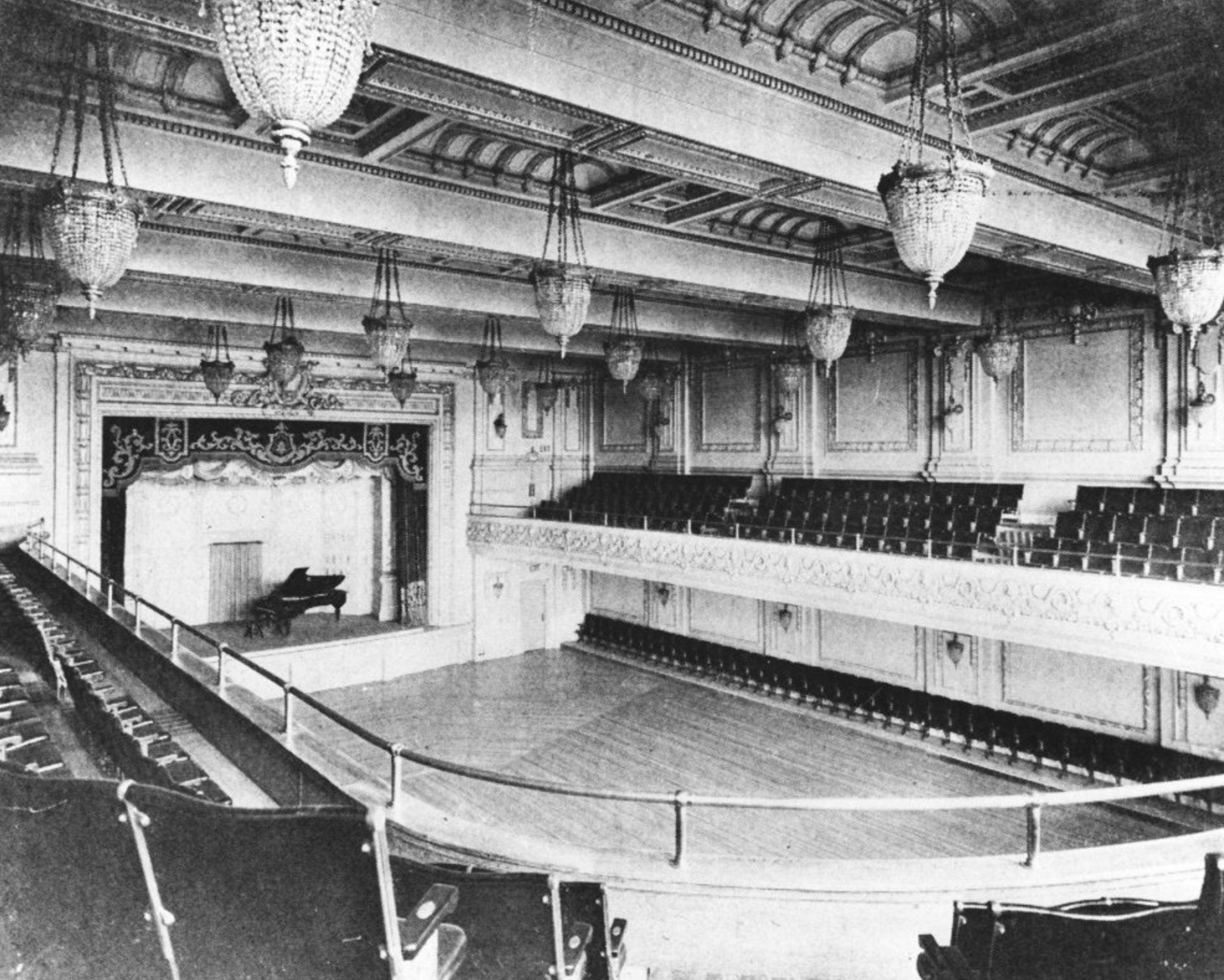 The image shows an elegant concert hall with a grand stage featuring a piano. Rows of empty seats fill the space, with ornate chandeliers hanging from the ceiling.