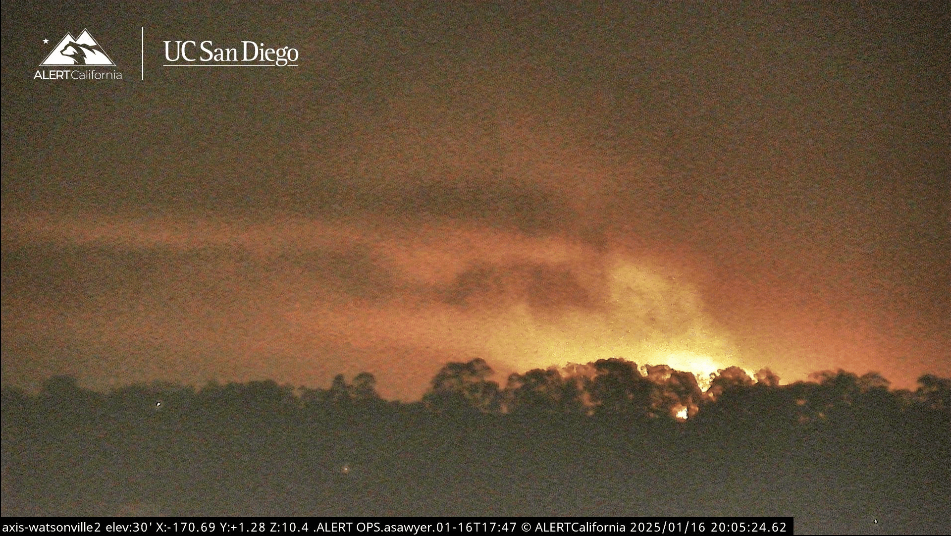 The image shows a wildfire with bright flames and smoke rising above a silhouette of trees under a night sky. It is branded with UC San Diego and ALERT California.