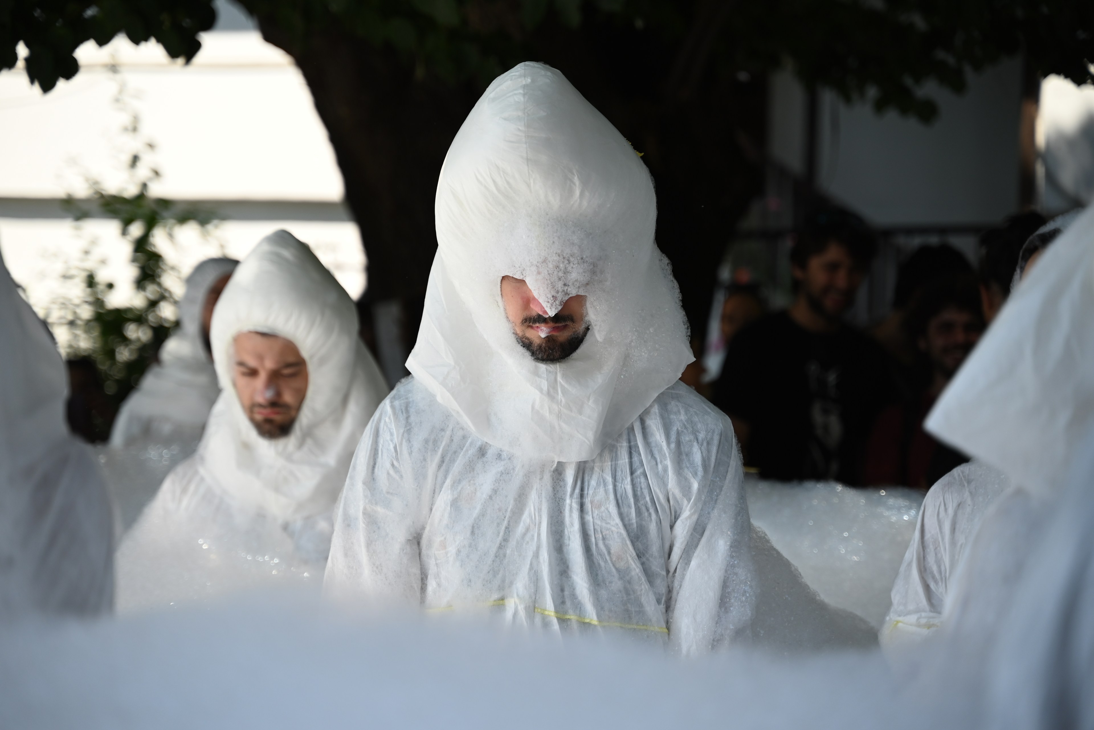 People in white protective suits stand in foam, their hoods inflated. The scene is outdoors with some people in the background.