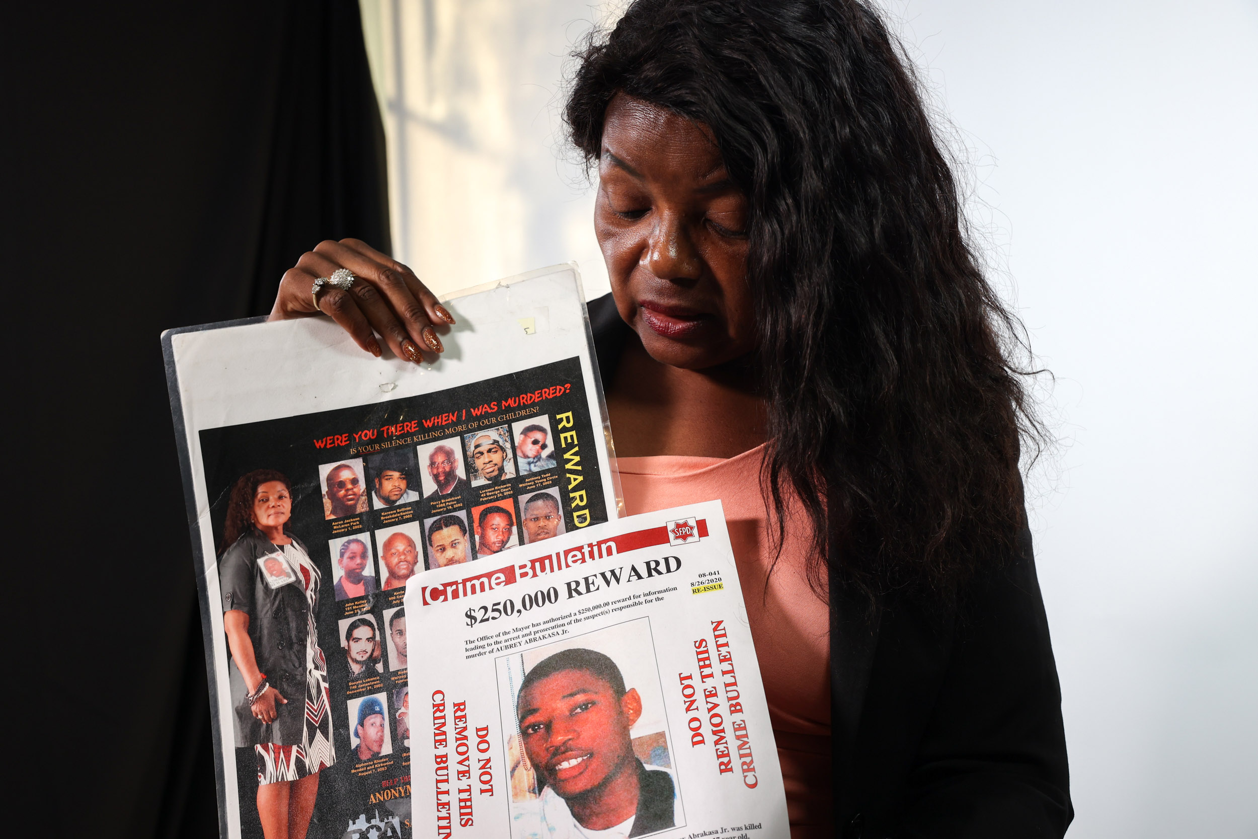 A woman with long hair holds two crime bulletin flyers, one offering a reward, featuring multiple photos of individuals in a somber and serious setting.