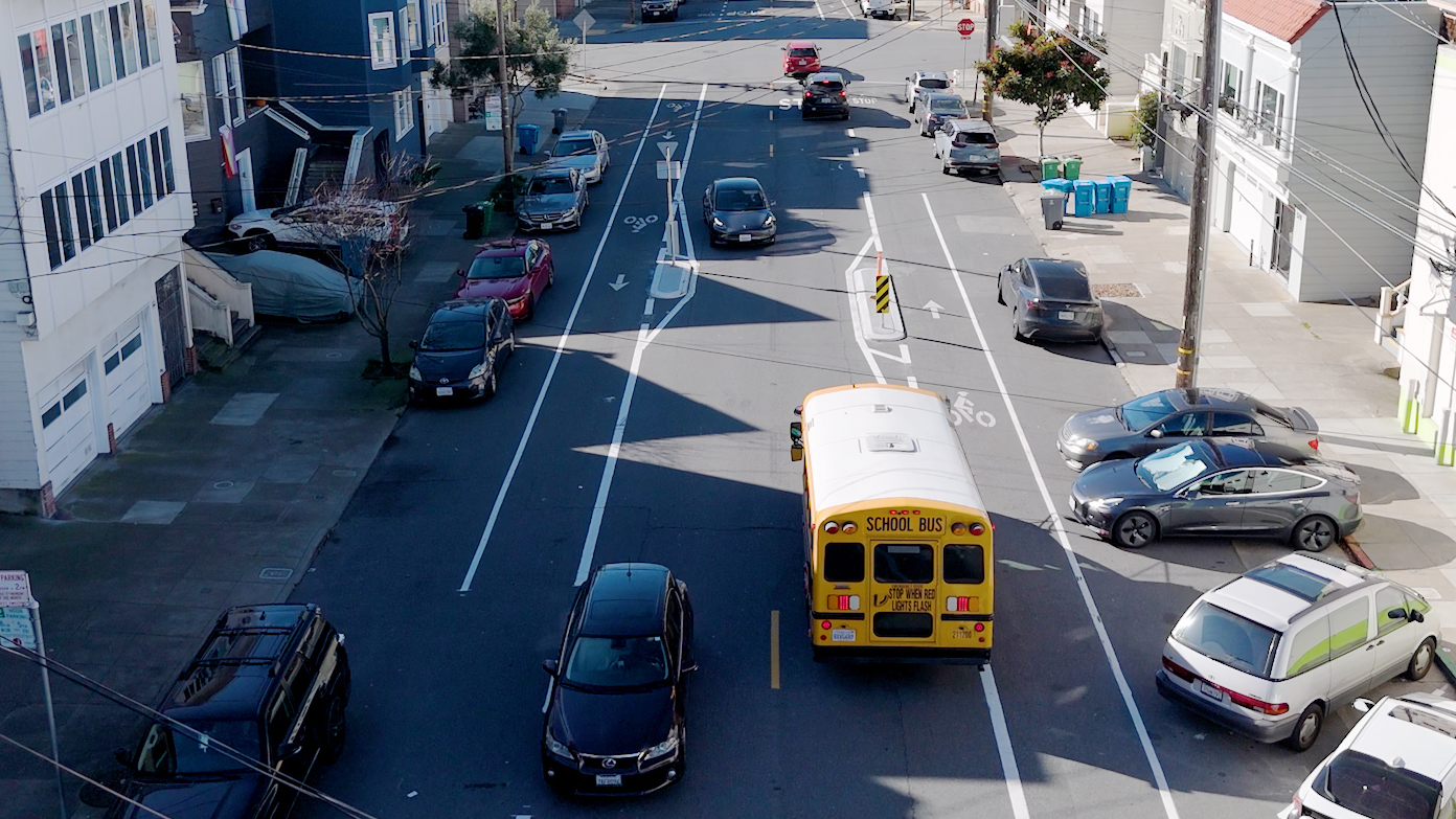 The image shows a suburban street with cars and a school bus. Vehicles are parked along the side, and there's a bicycle lane marked on the road.