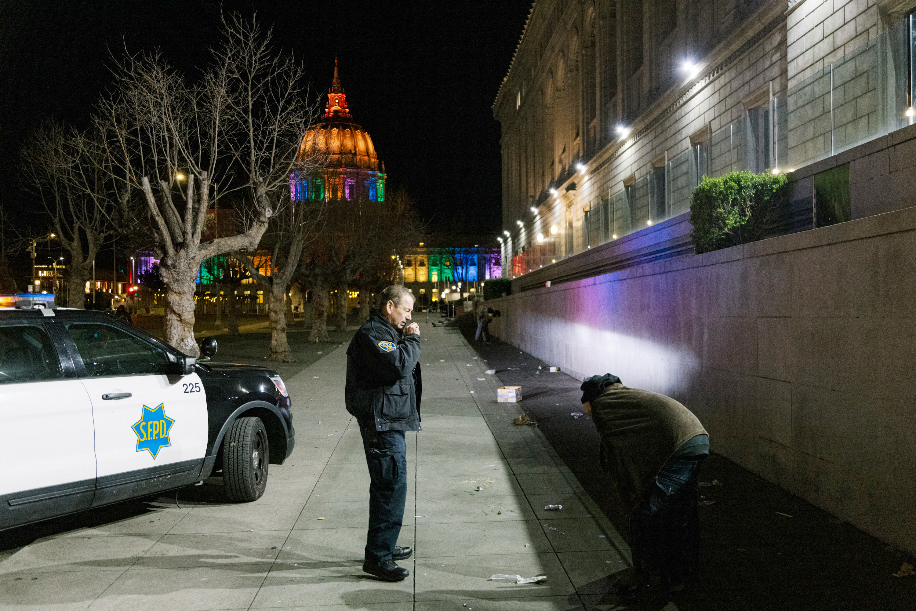 A police officer stands near a squad car outside a building. The building and nearby trees are illuminated, creating a colorful night scene.