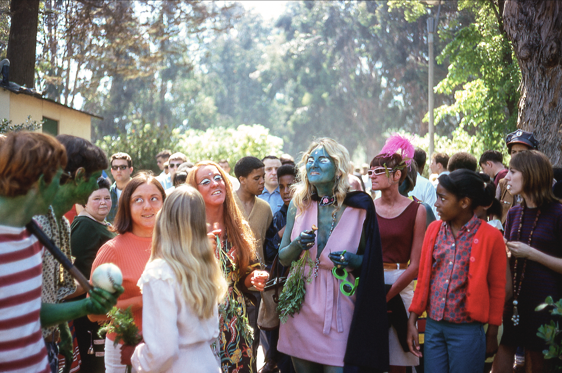 A diverse crowd gathers outdoors, with individuals in colorful costumes, face paint, and playful outfits, chatting and enjoying a vibrant, sunny atmosphere.