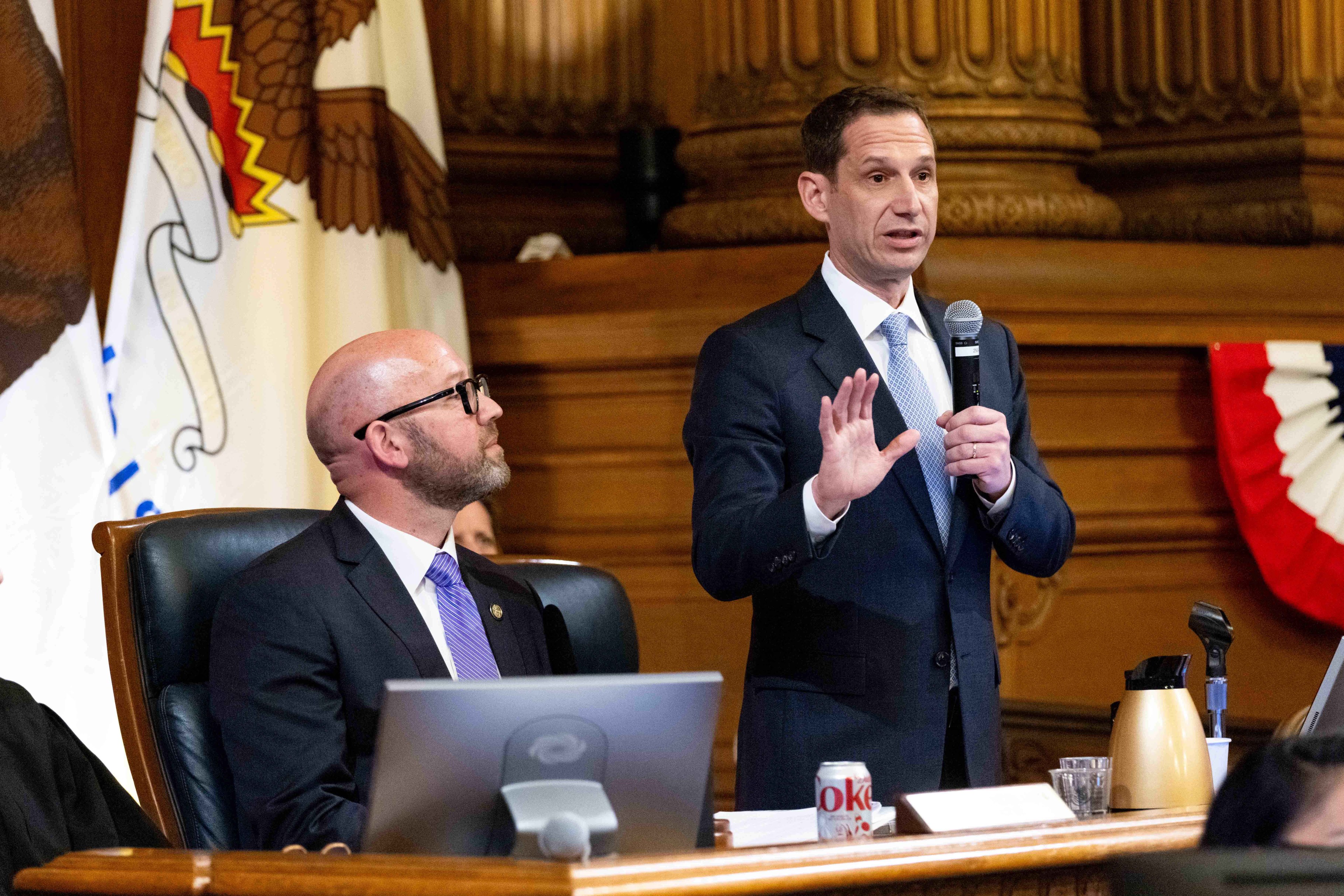 A man in a suit speaks into a microphone, gesturing, while another man in a suit sits at a desk with a laptop and Diet Coke in front of him. There's a US flag behind them.