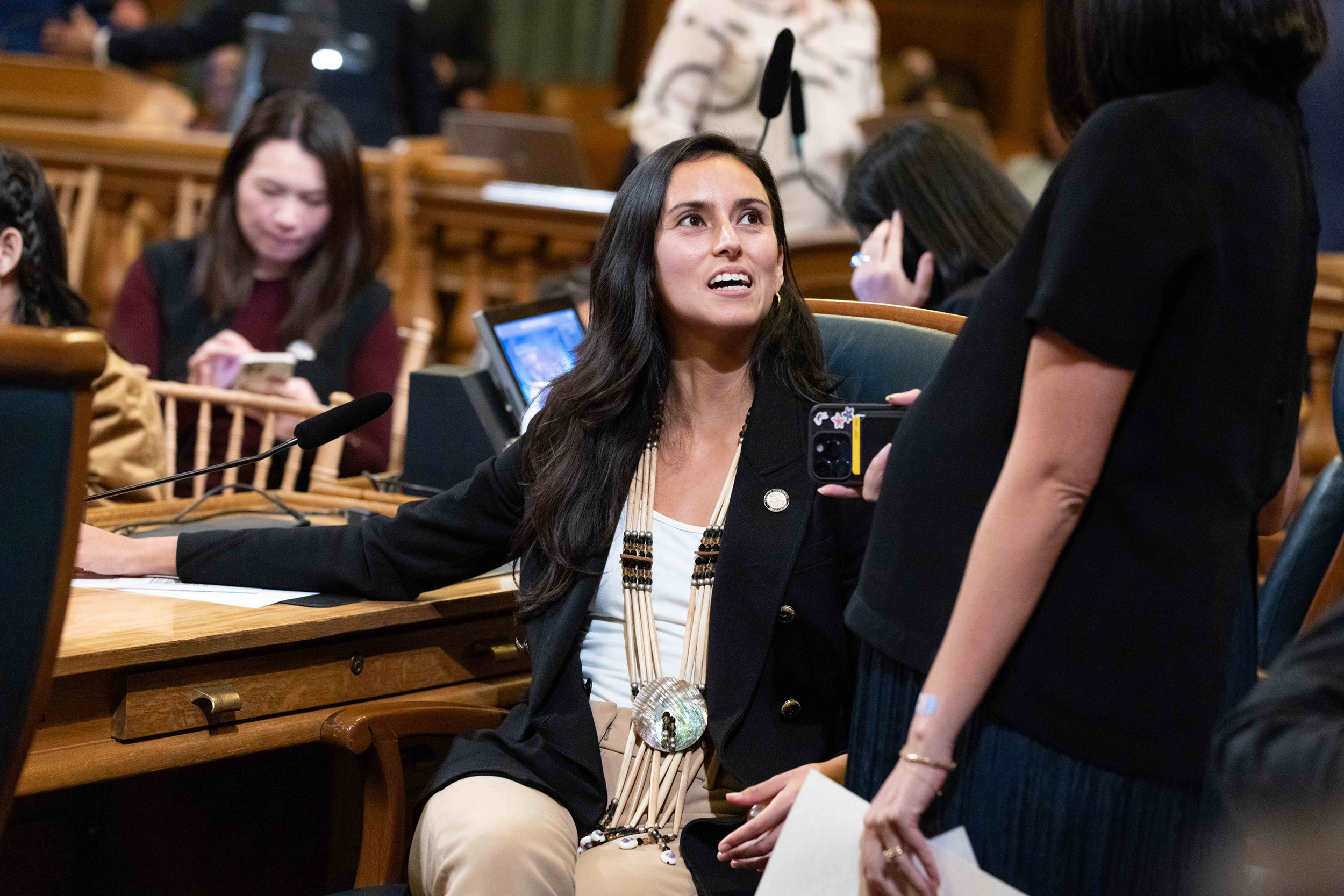 A woman in a formal setting is seated and speaking to another standing woman holding a phone. She wears a statement necklace and there are people working in the background.