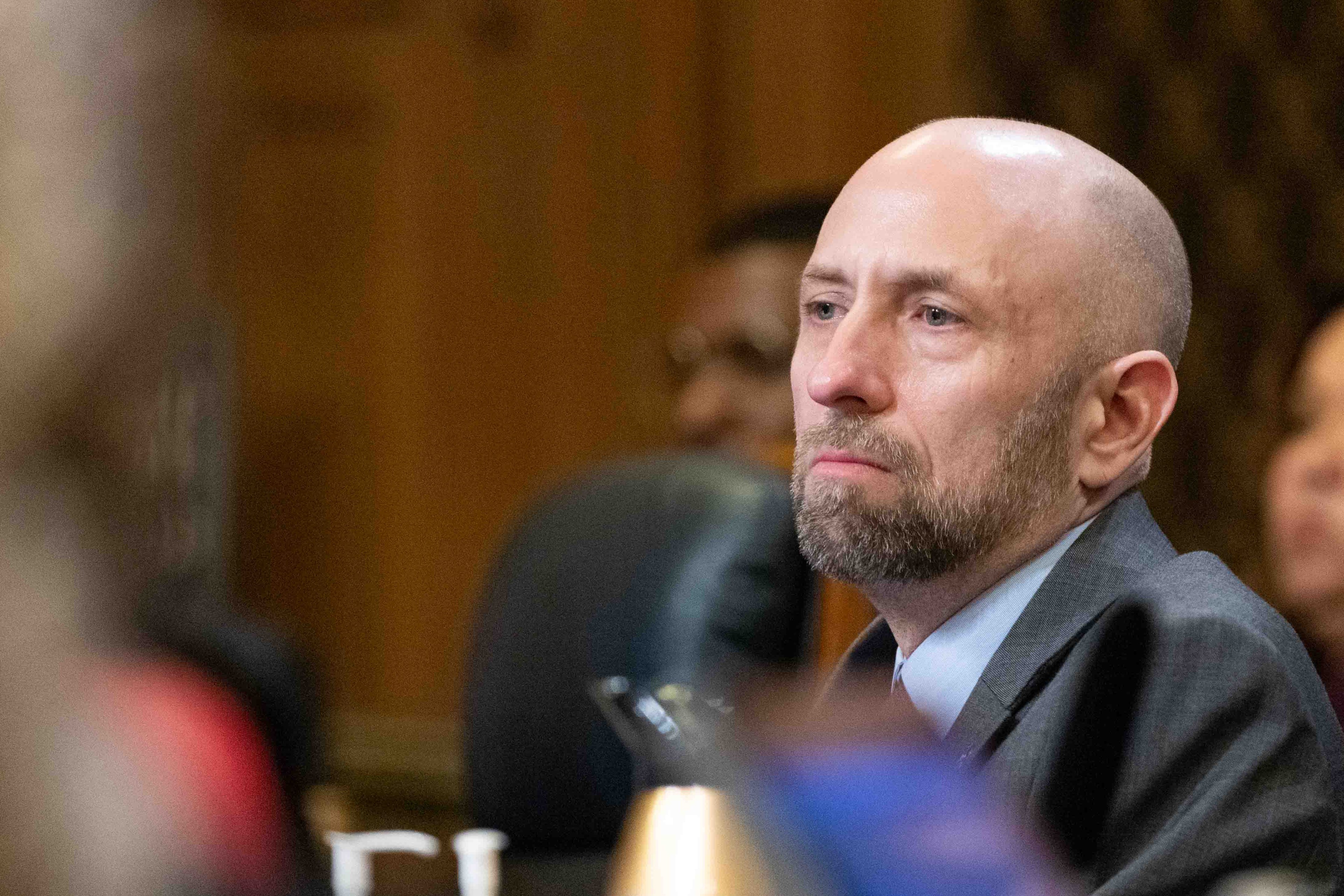 A man with a bald head and beard is sitting attentively in a formal setting, wearing a suit. The background is blurred, suggesting he is in a meeting or conference.