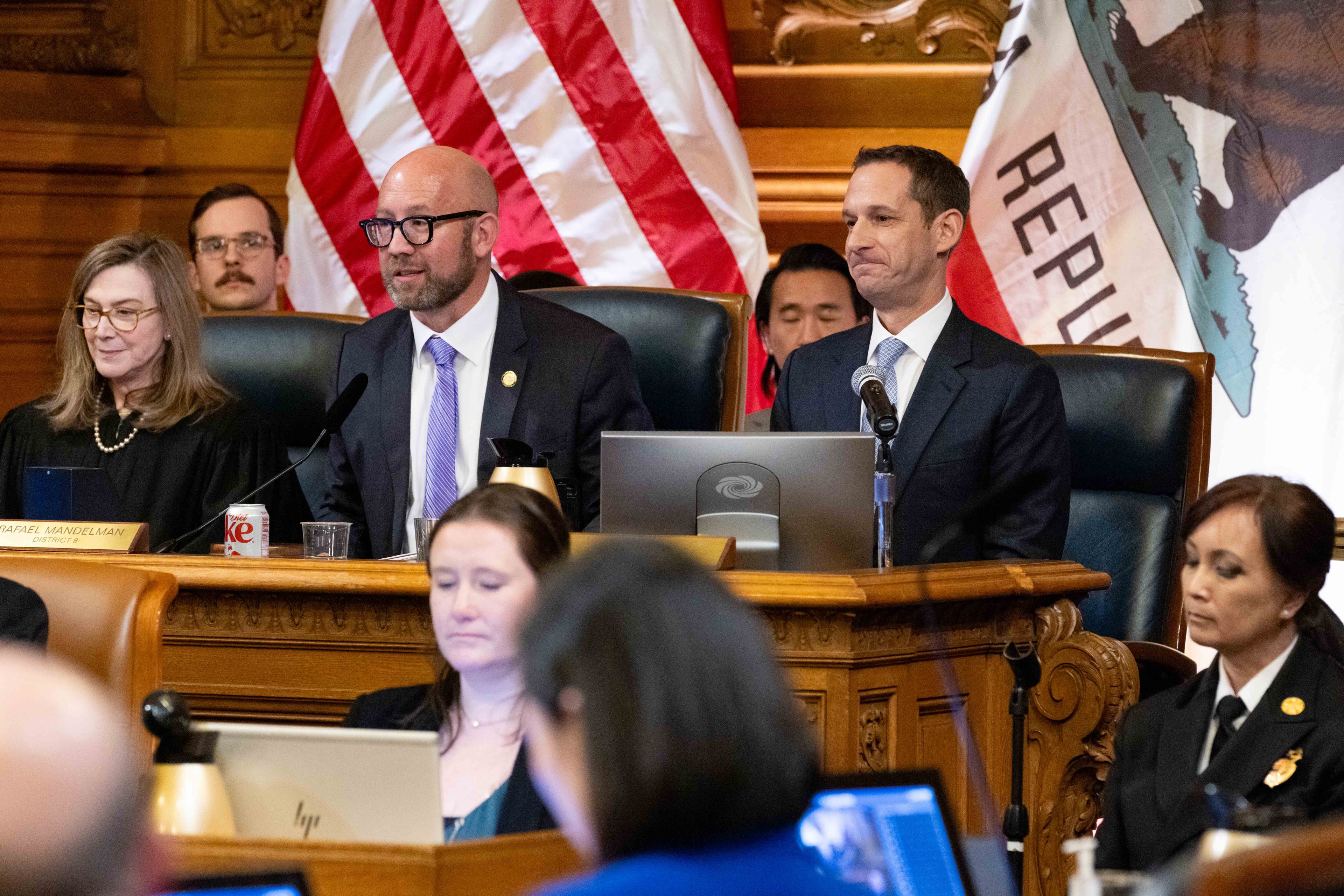 Several people are seated in a formal setting with microphones and flags in the background, suggesting a government or official meeting. They appear to be engaged in discussion.