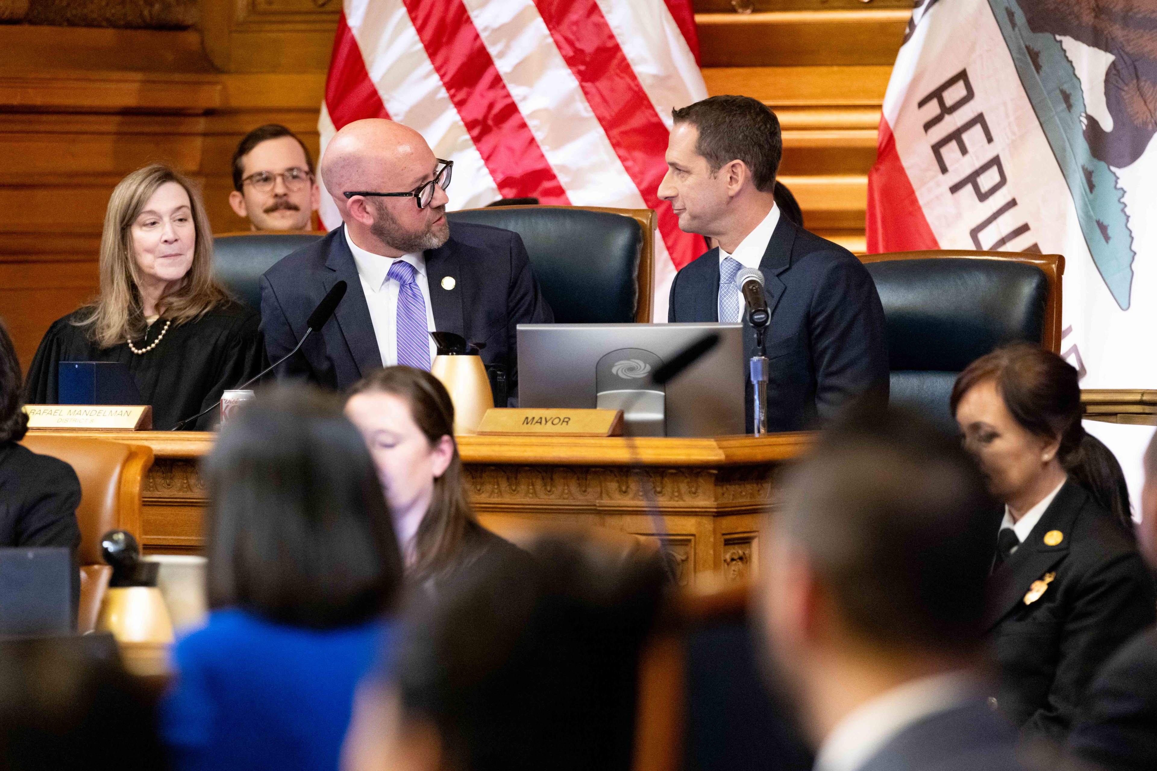 Several people are seated at a large, ornate desk in a formal setting, with two men in suits engaged in conversation. American and California flags are visible.