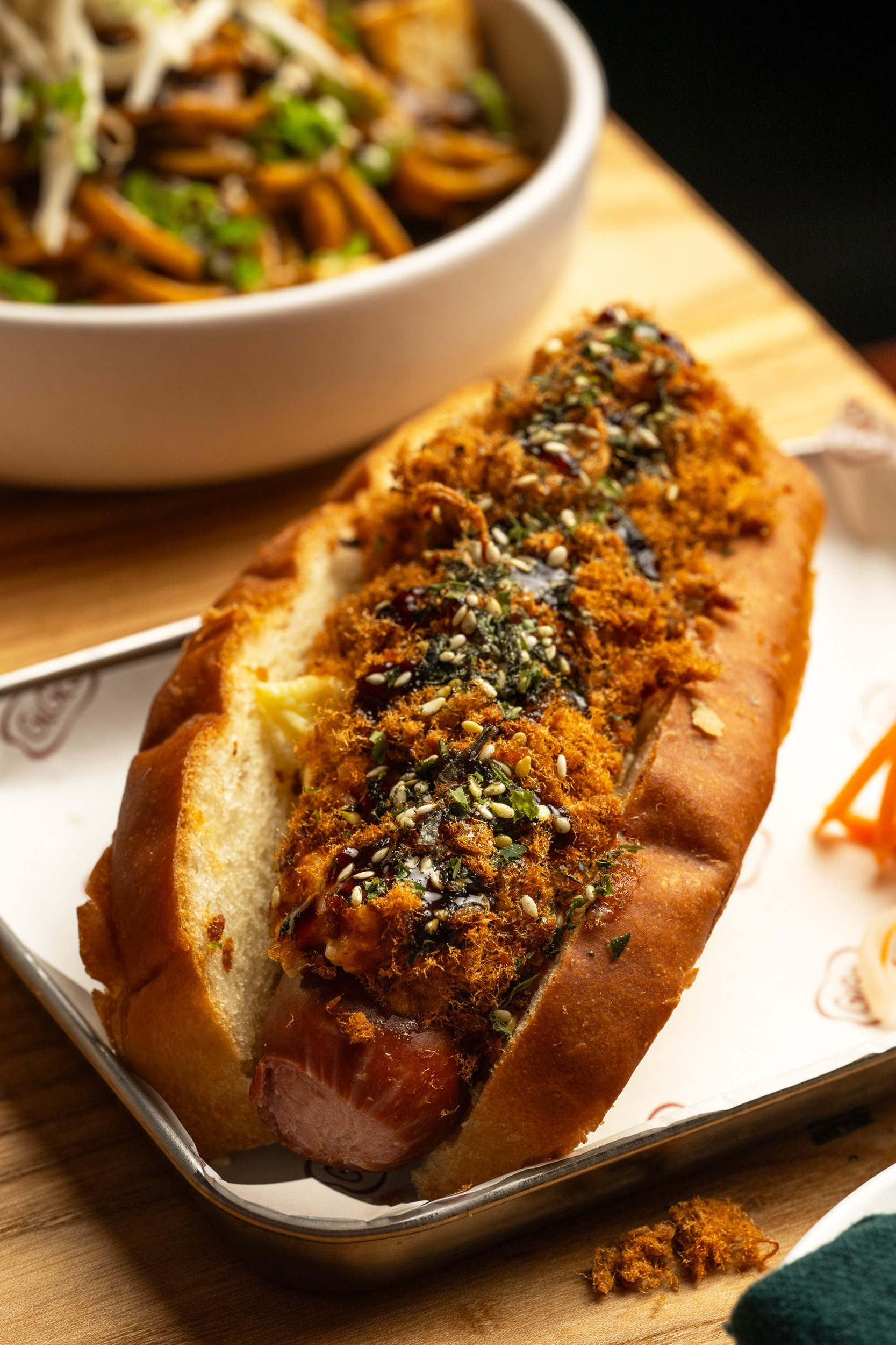 A hot dog with savory toppings like shredded pork floss and sesame seeds rests in a toasted bun on a tray. A bowl of noodle salad is in the background.