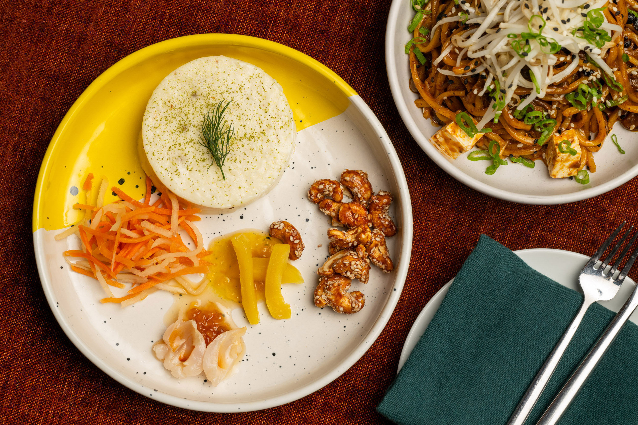 A yellow and white plate holds a soft cheese topped with herbs, and small servings of pickled veggies and nuts. Nearby is a noodle dish with tofu and greens.