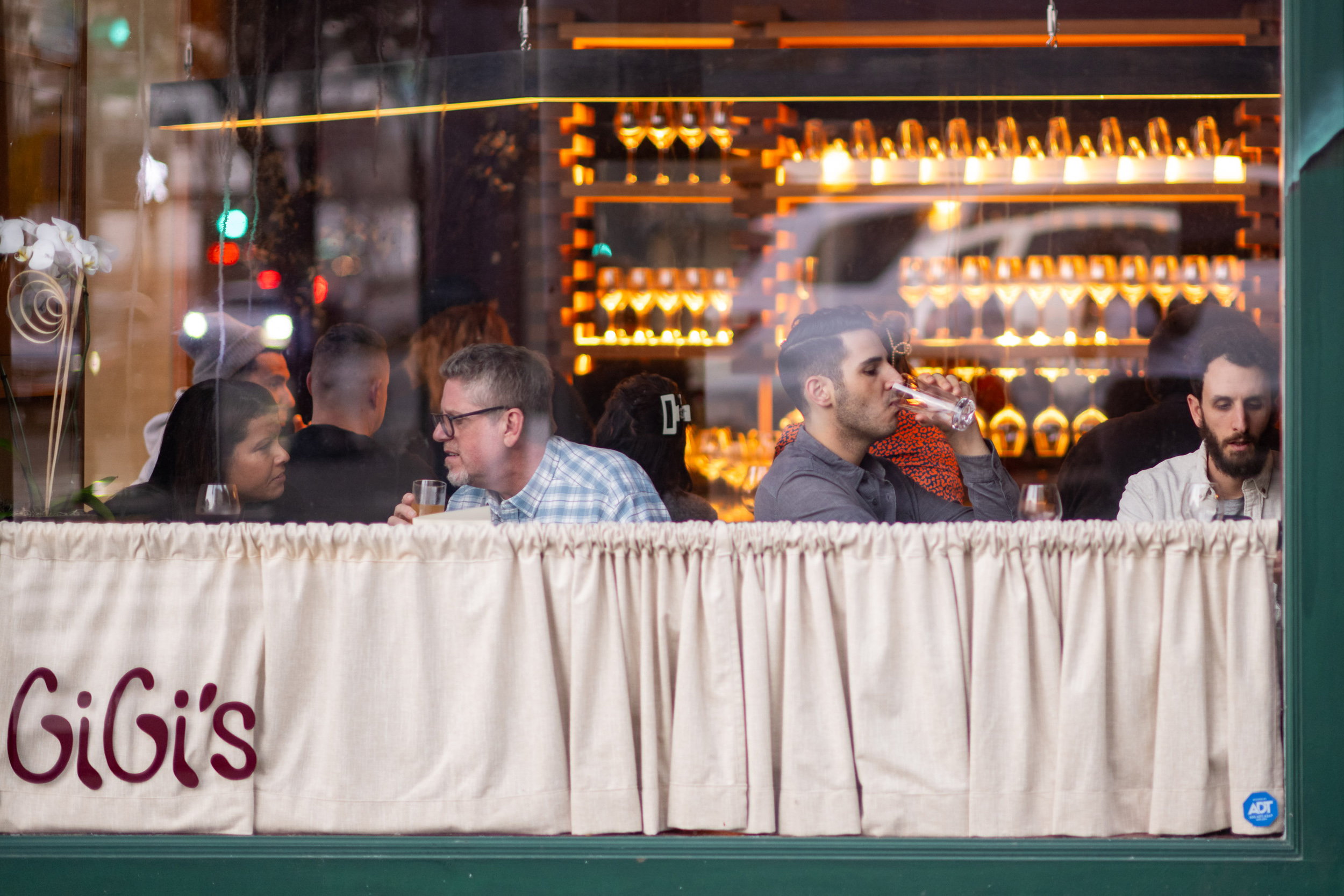 People are sitting at a window-side table in a restaurant called GiGi's, having drinks. Behind them is a wall with neatly arranged wine glasses.