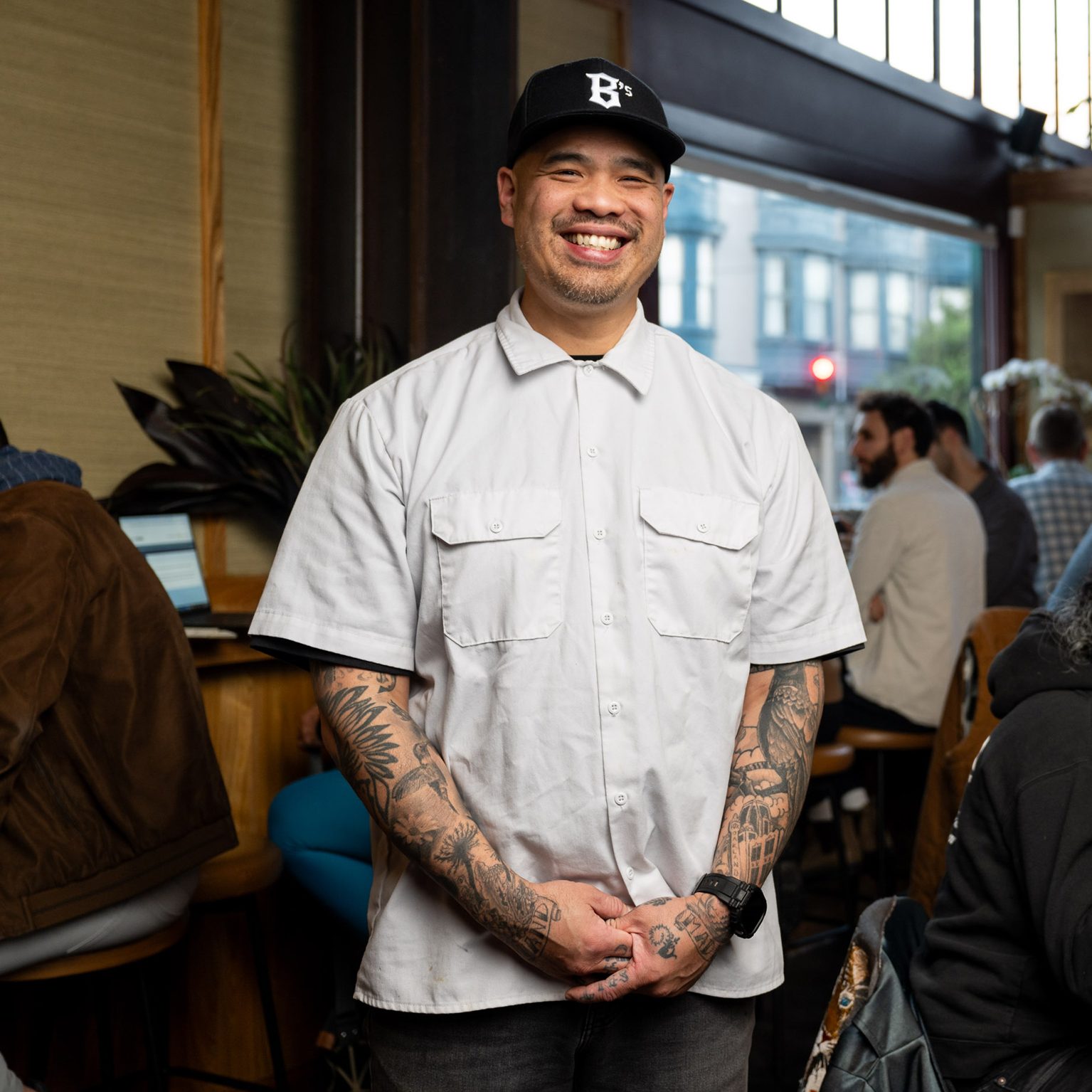 A smiling man wearing a white shirt and cap stands in a bustling restaurant. People sit at a bar, eating and conversing, with a window showing a street view.