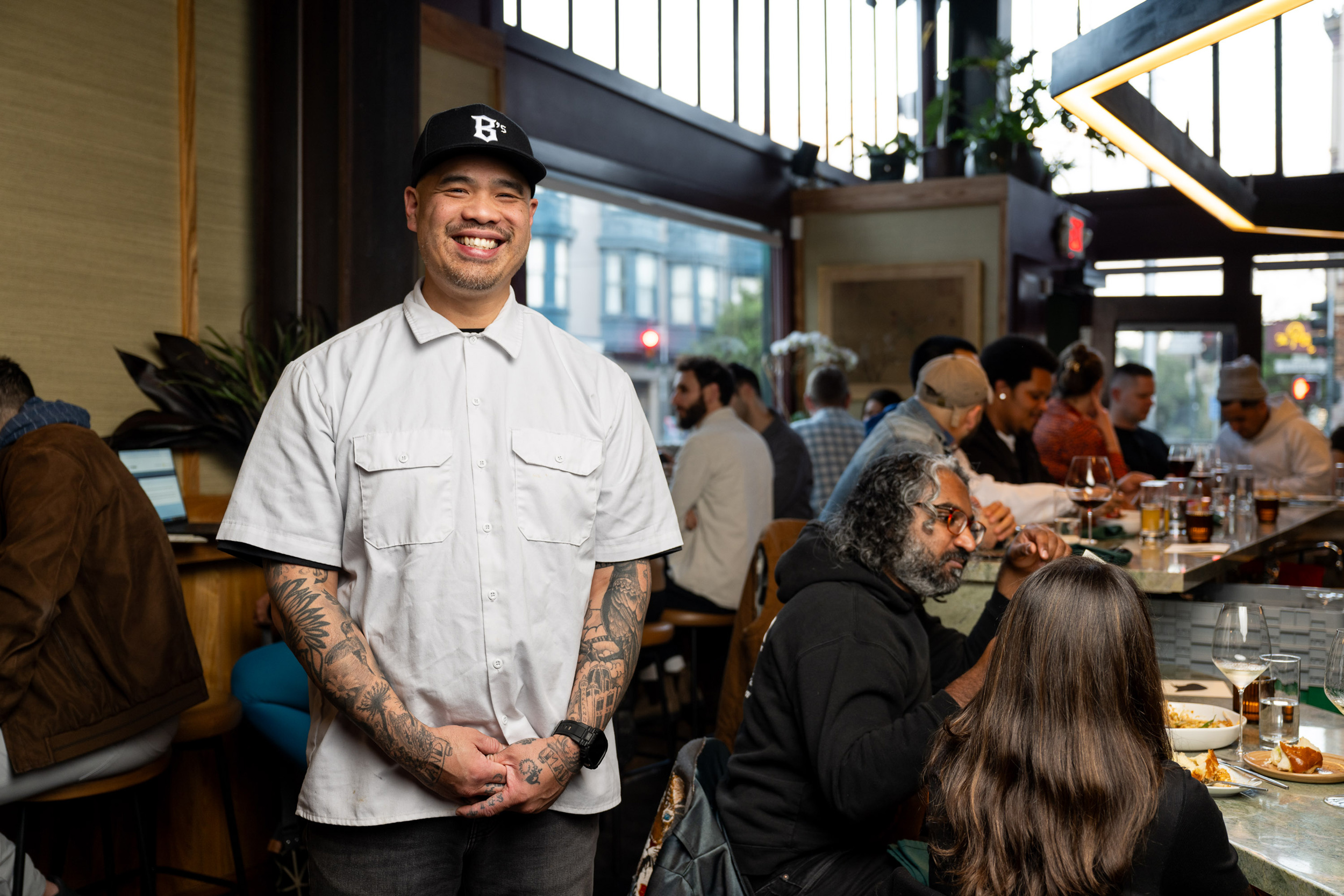 A smiling man wearing a white shirt and cap stands in a bustling restaurant. People sit at a bar, eating and conversing, with a window showing a street view.