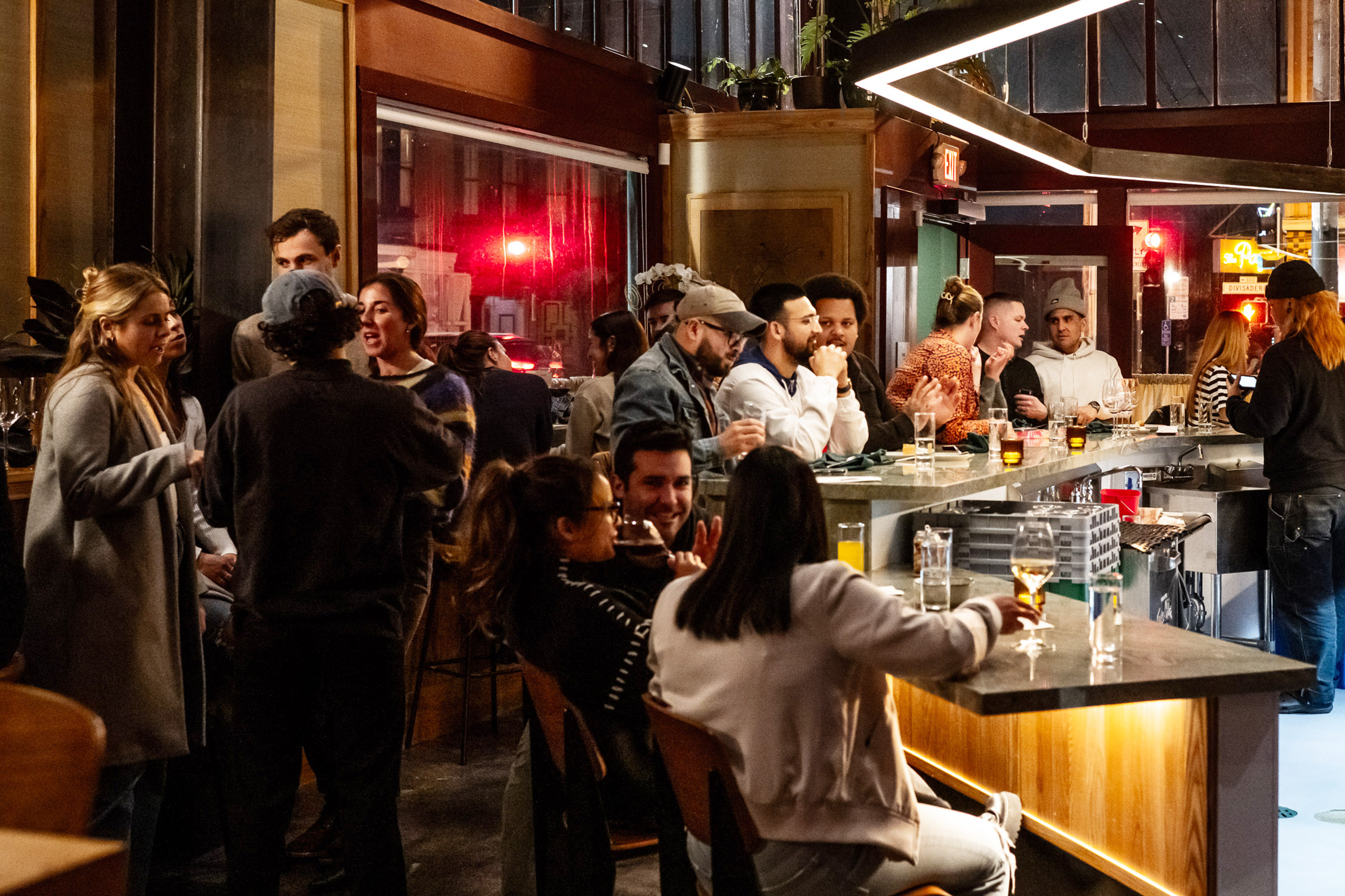 A lively bar scene shows people mingling and chatting around a long counter. Drinks are on the tables, and the atmosphere is warm and social with dim lighting.