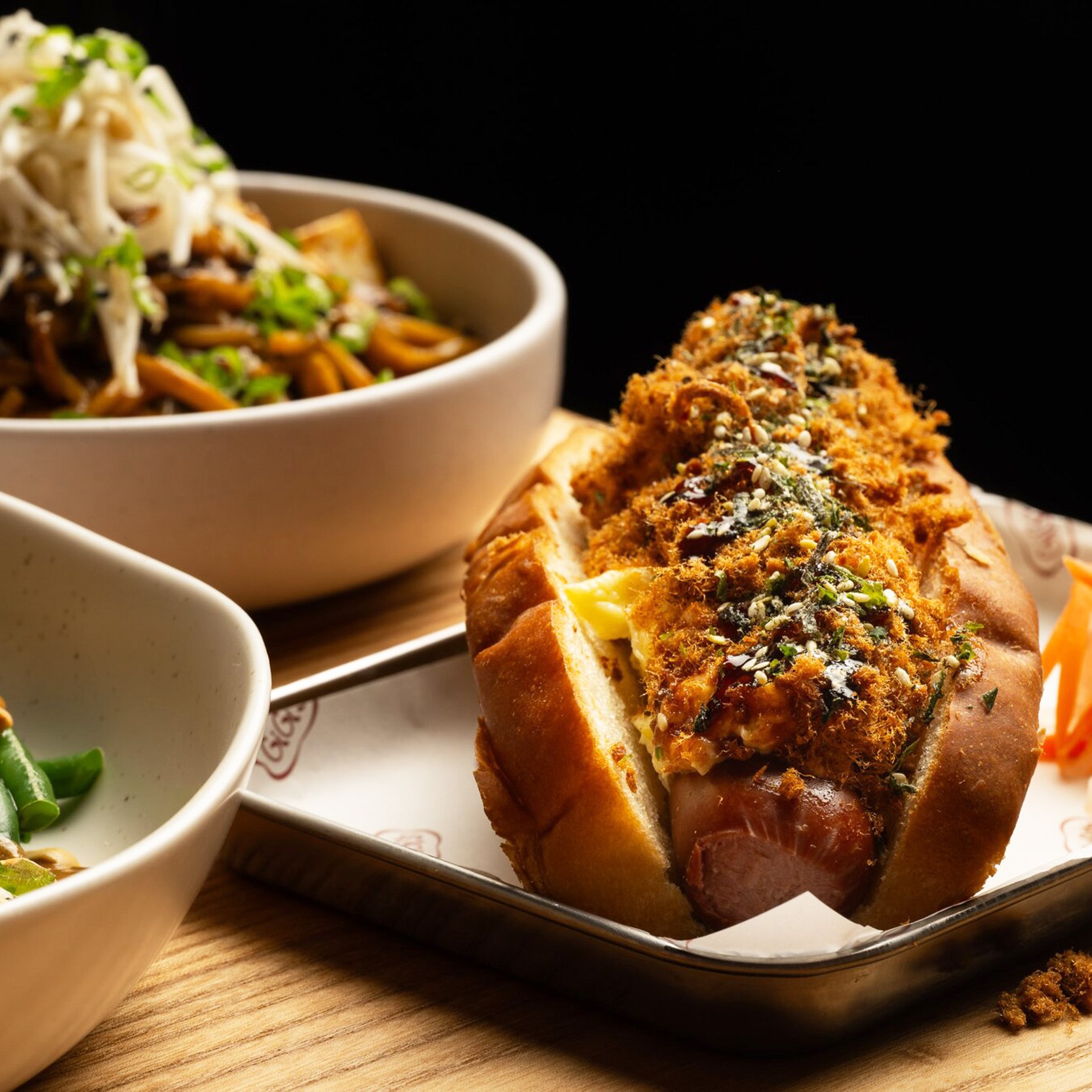 The image shows a plated hot dog topped with herbs and crispy bits, alongside bowls of noodles with sprouts and salad with radish and green beans.