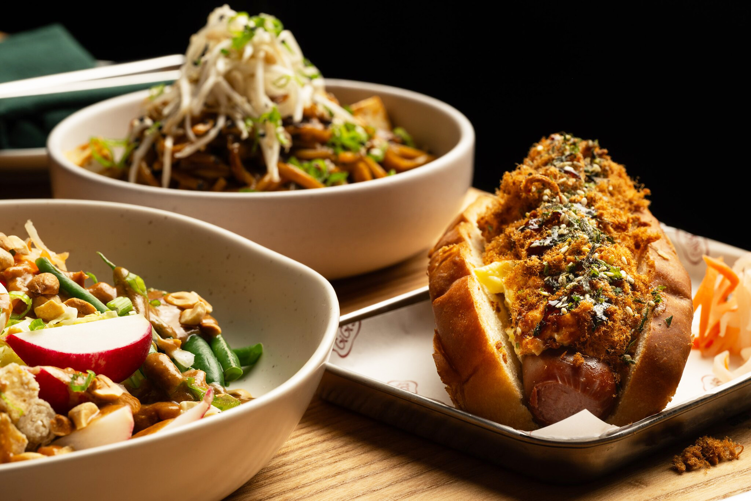 The image shows a plated hot dog topped with herbs and crispy bits, alongside bowls of noodles with sprouts and salad with radish and green beans.