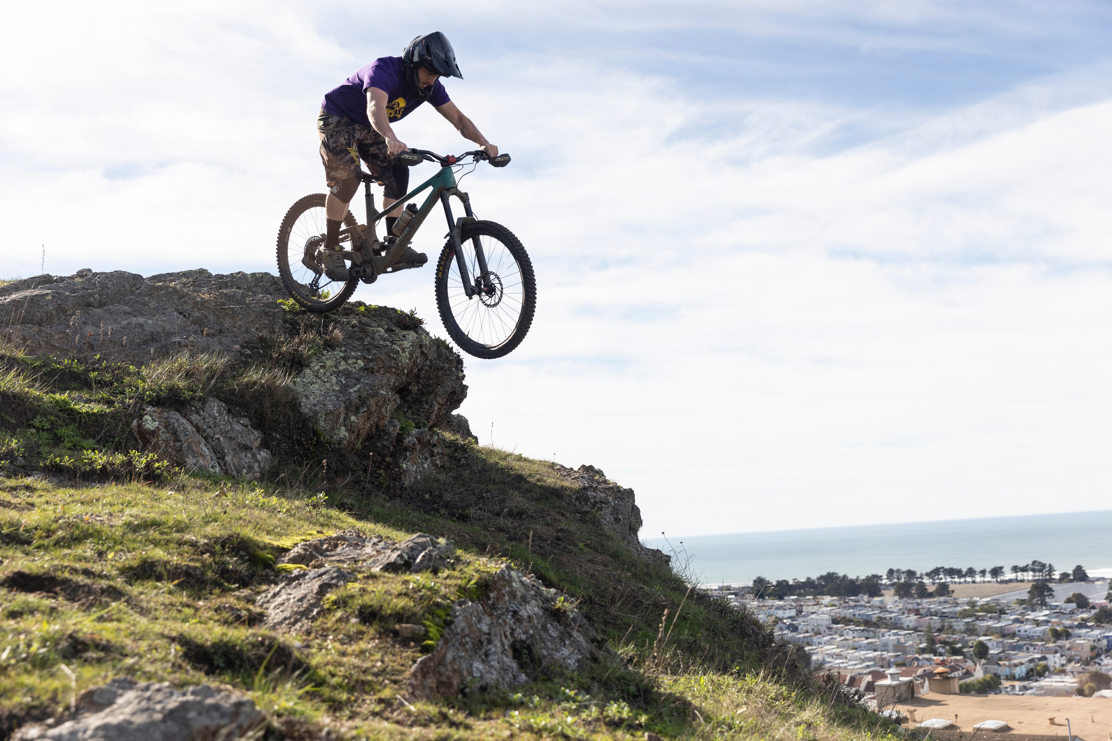 A cyclist in a helmet descends a rocky hill on a mountain bike. The backdrop features a distant coastline and a town under a partly cloudy sky.