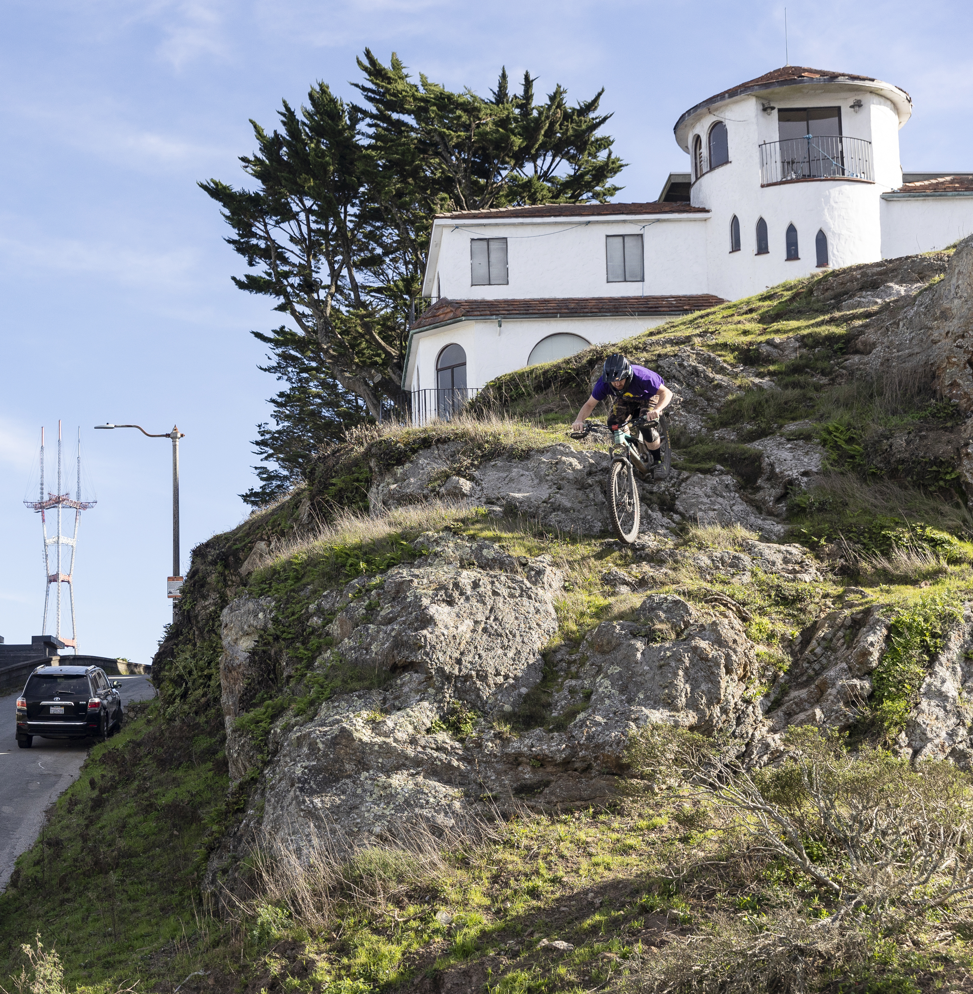 A cyclist in a purple shirt rides downhill on a rocky slope. In the background, there's a white house with arched windows and a tall structure to the left.