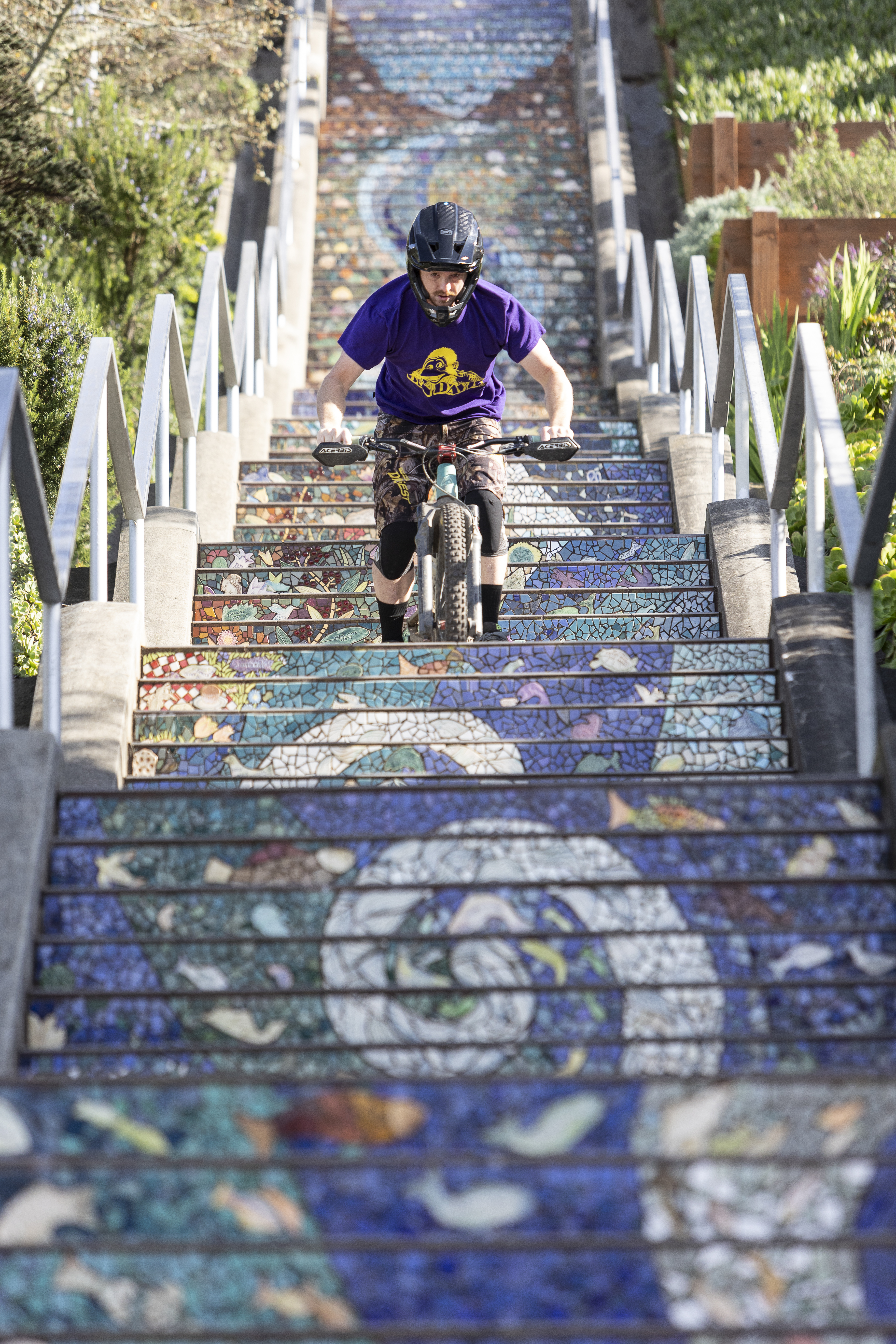 A person in a purple shirt and helmet rides a bike down colorful mosaic stairs, surrounded by greenery and artistic railings.