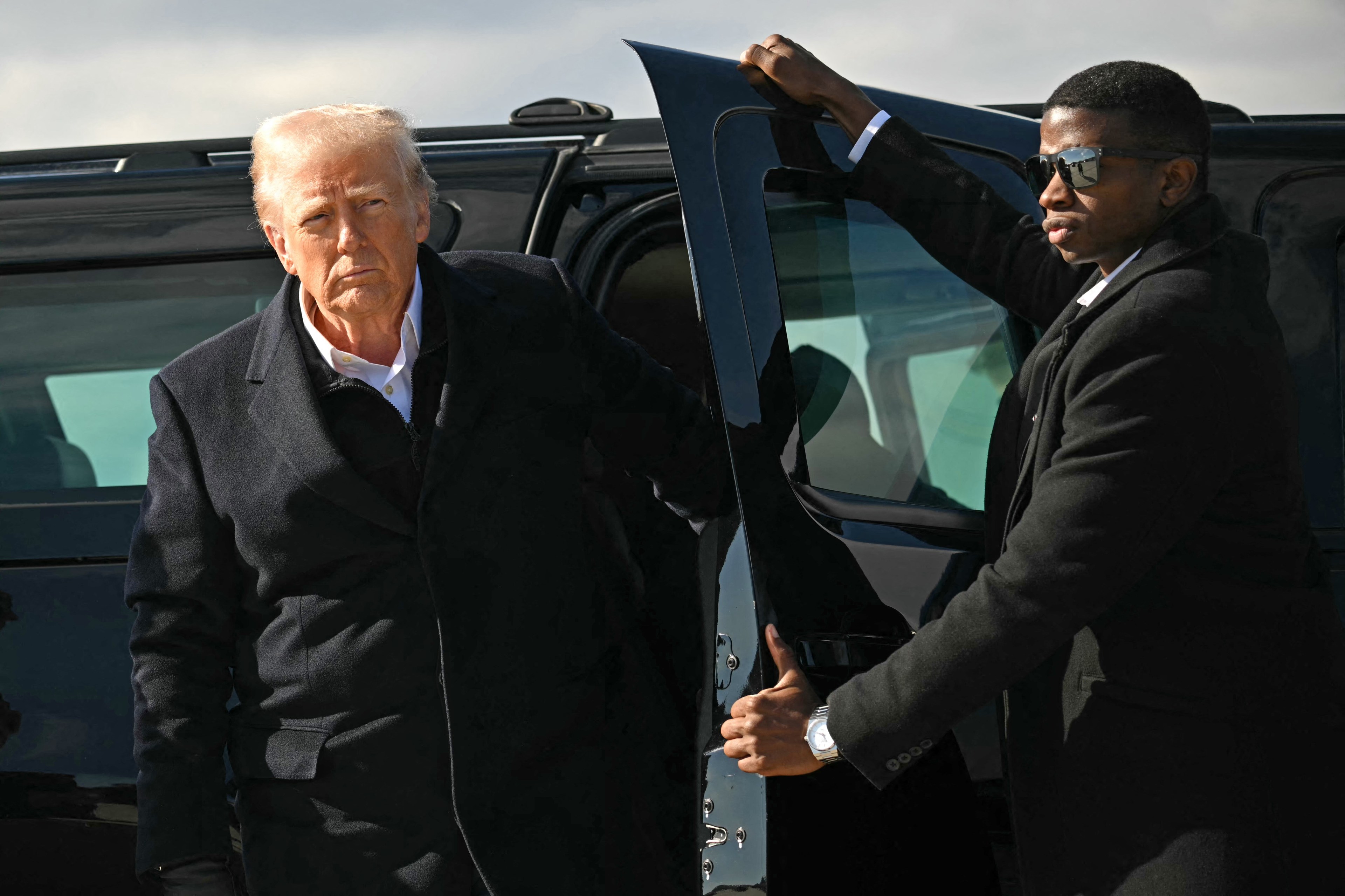 A man in a dark coat exits a black vehicle, assisted by another man in sunglasses holding the door. They're outdoors, with a clear sky in the background.