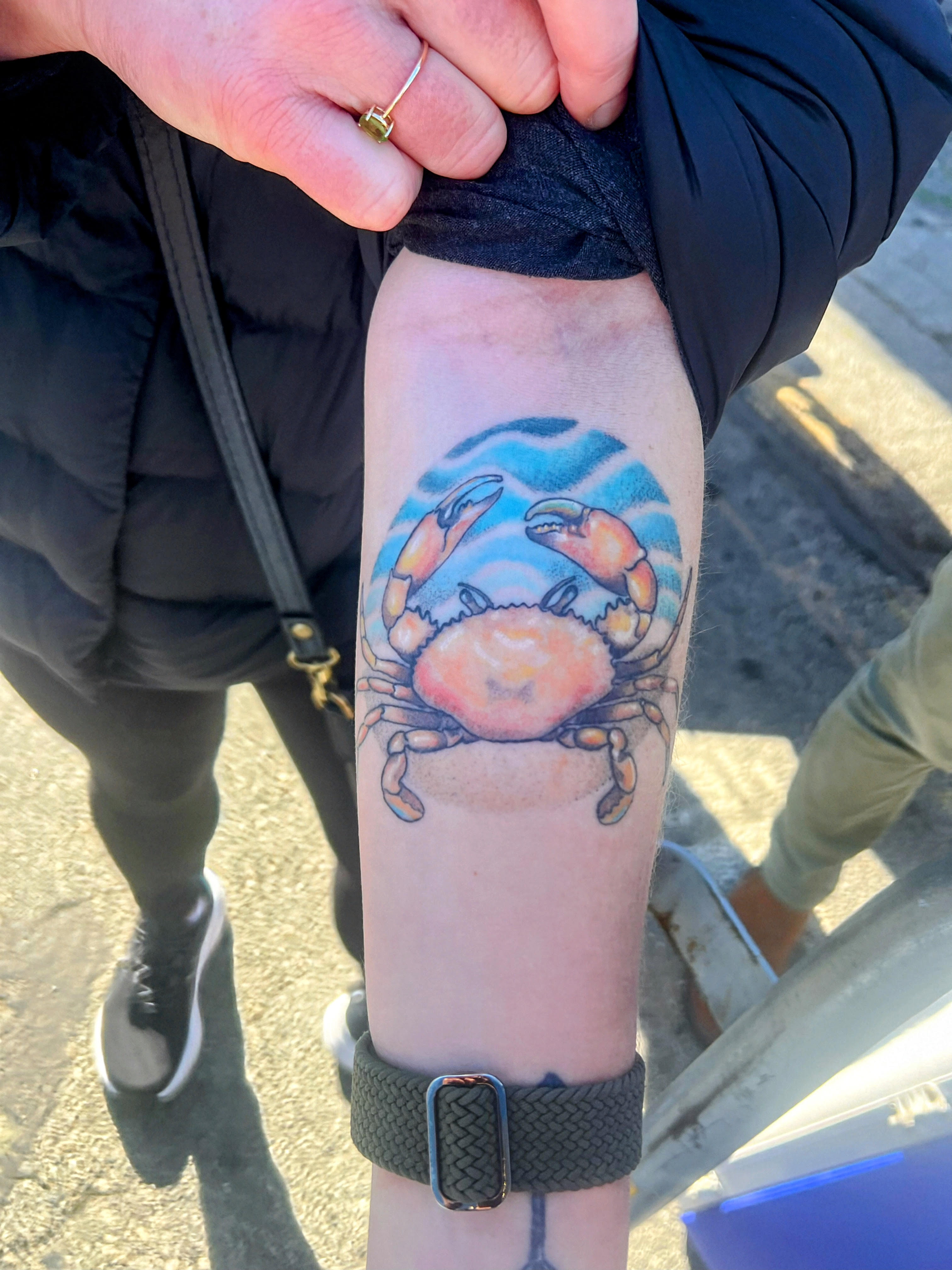 The image shows an arm tattoo of a colorful crab with blue waves in the background. A hand with a ring holds the sleeve up.