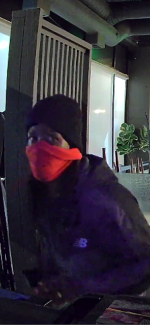 A person is indoors, wearing a black beanie and a red face mask. They're dressed in a dark jacket, standing near a modern interior wall with plants visible.