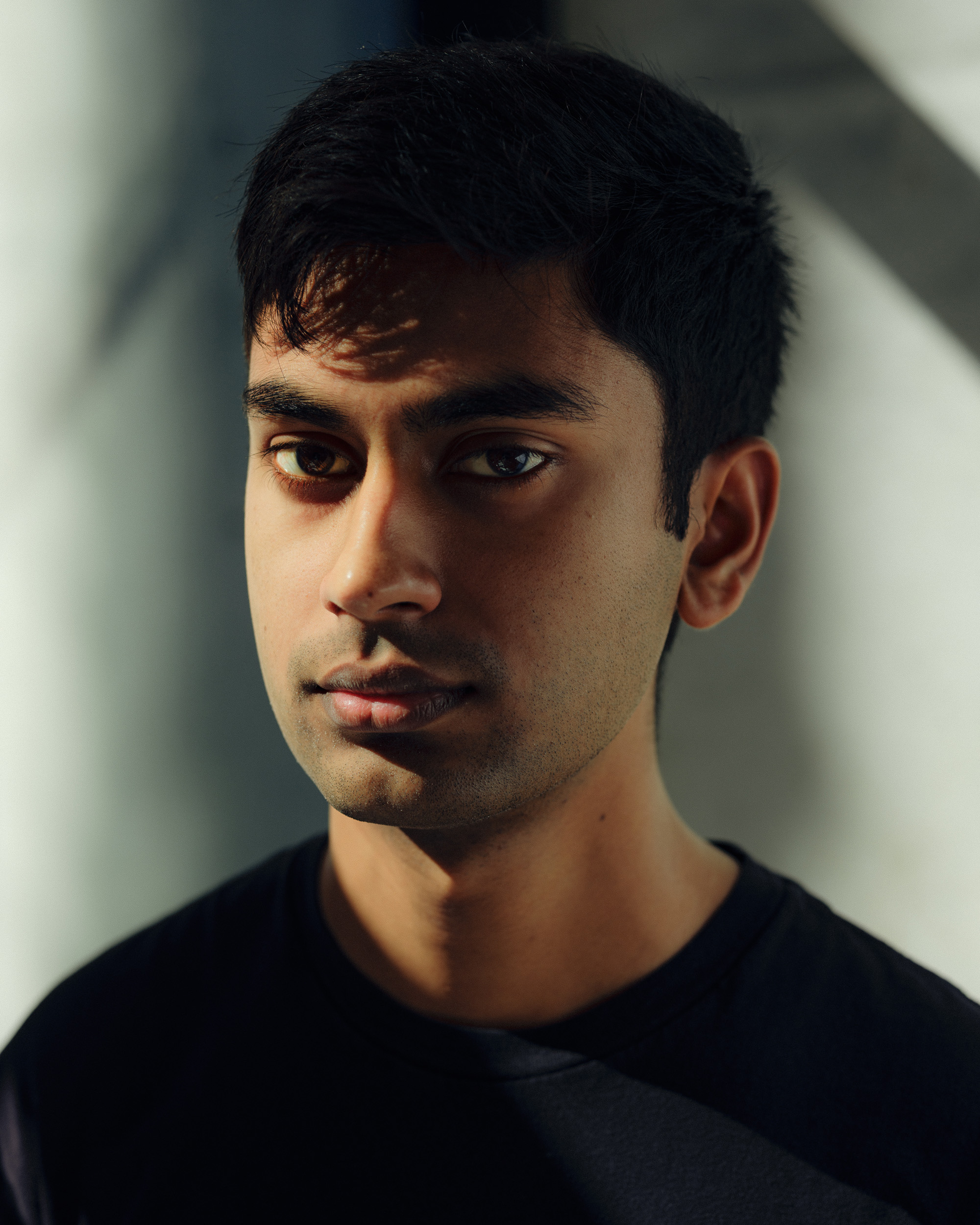 Suchir Balaji looks directly at the camera. The lighting casts soft shadows on his face, creating a dramatic effect. He wears a dark shirt.