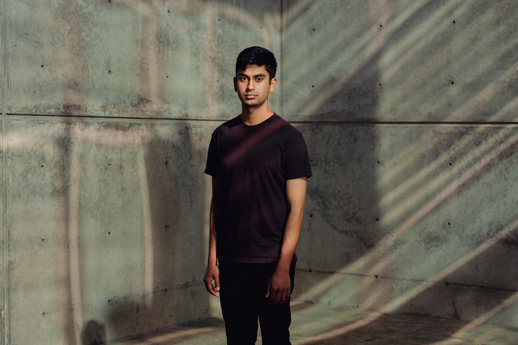 Suchir Balaji stands in a concrete-walled space, illuminated by diagonal light and shadow patterns. He wears a black shirt, dark pants, and white sneakers.