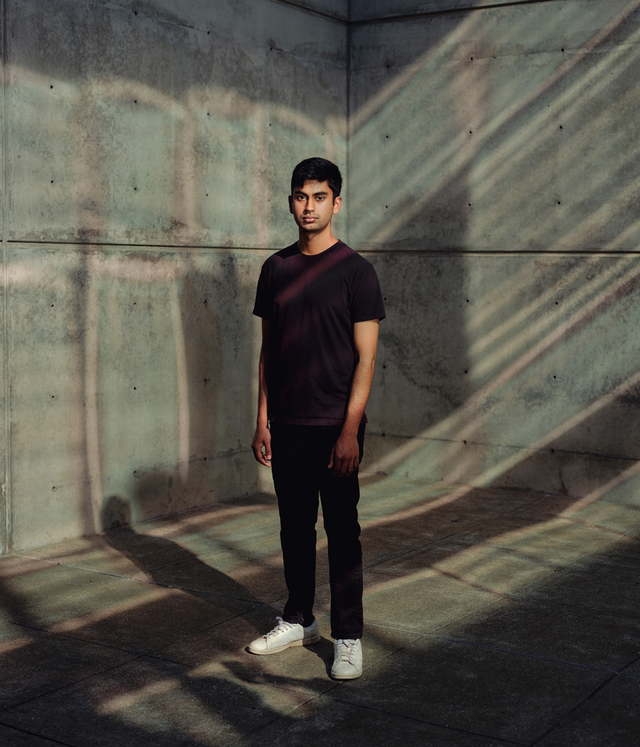 Suchir Balaji stands in a concrete-walled space, illuminated by diagonal light and shadow patterns. He wears a black shirt, dark pants, and white sneakers.