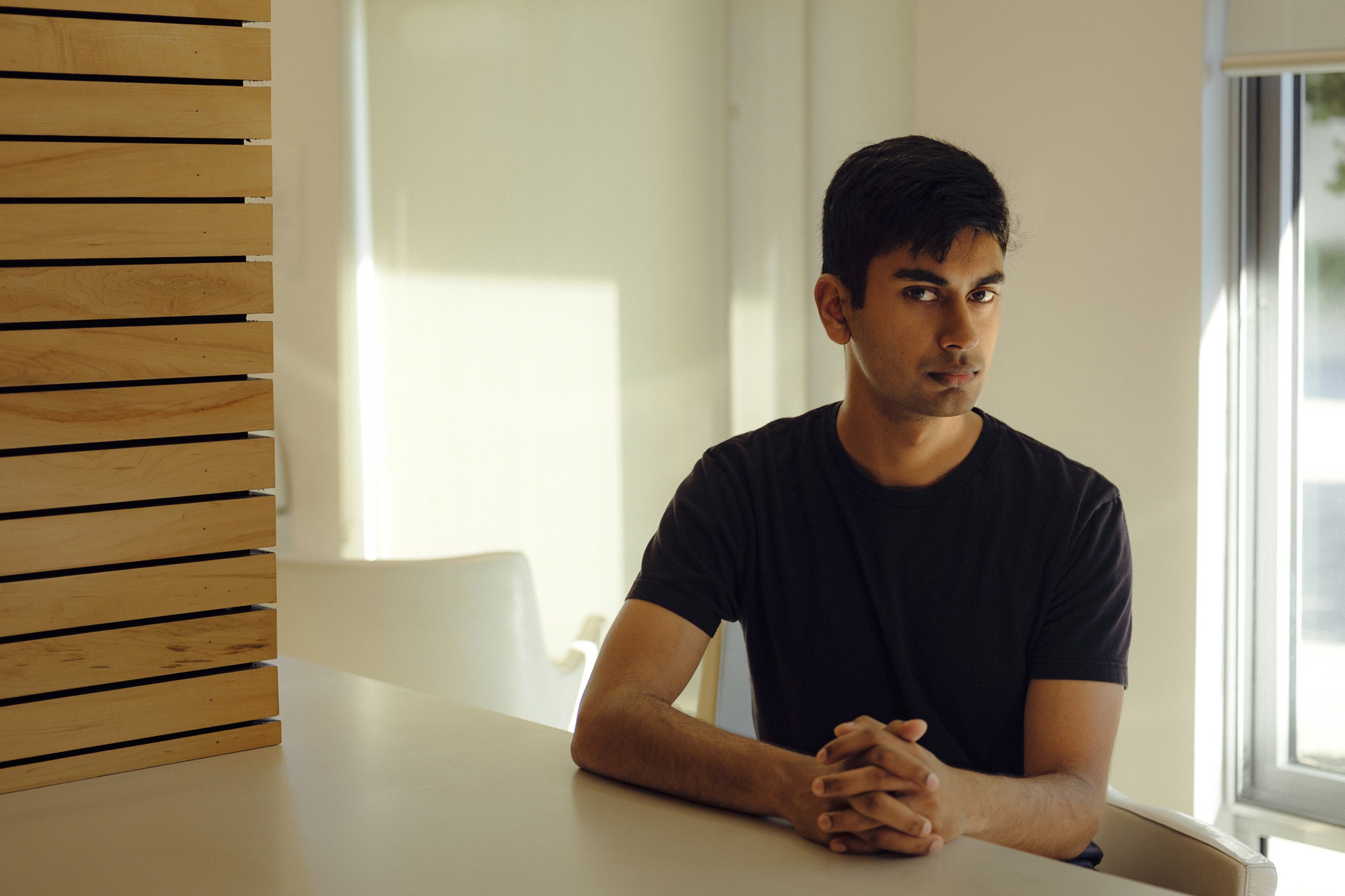 A person in a black shirt sits at a table with hands clasped. The setting is a well-lit room with a wooden panel wall and a window letting in natural light.